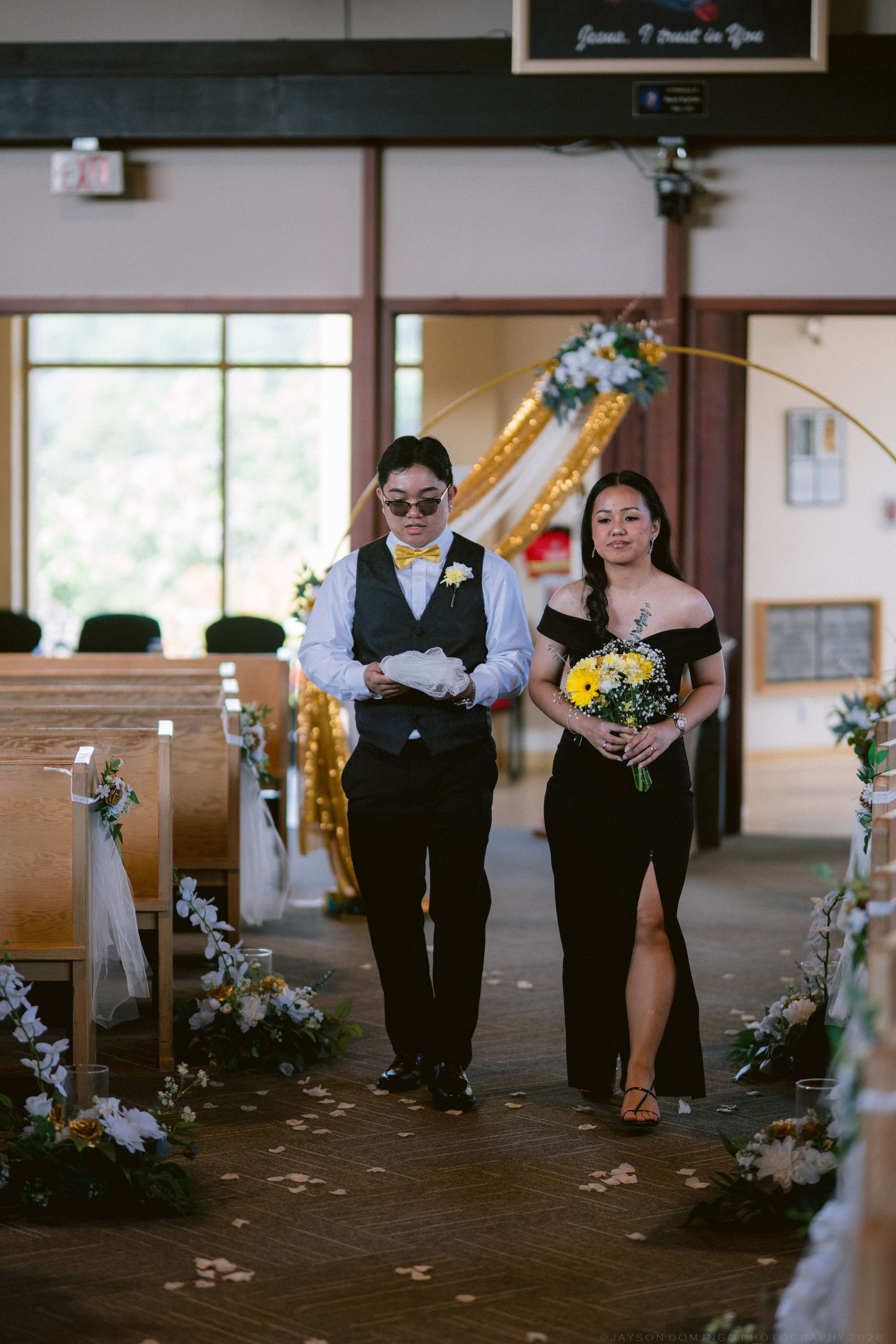 Groomsman in black vest escorts bridesmaid in black dress down flower-lined church aisle during wedding.