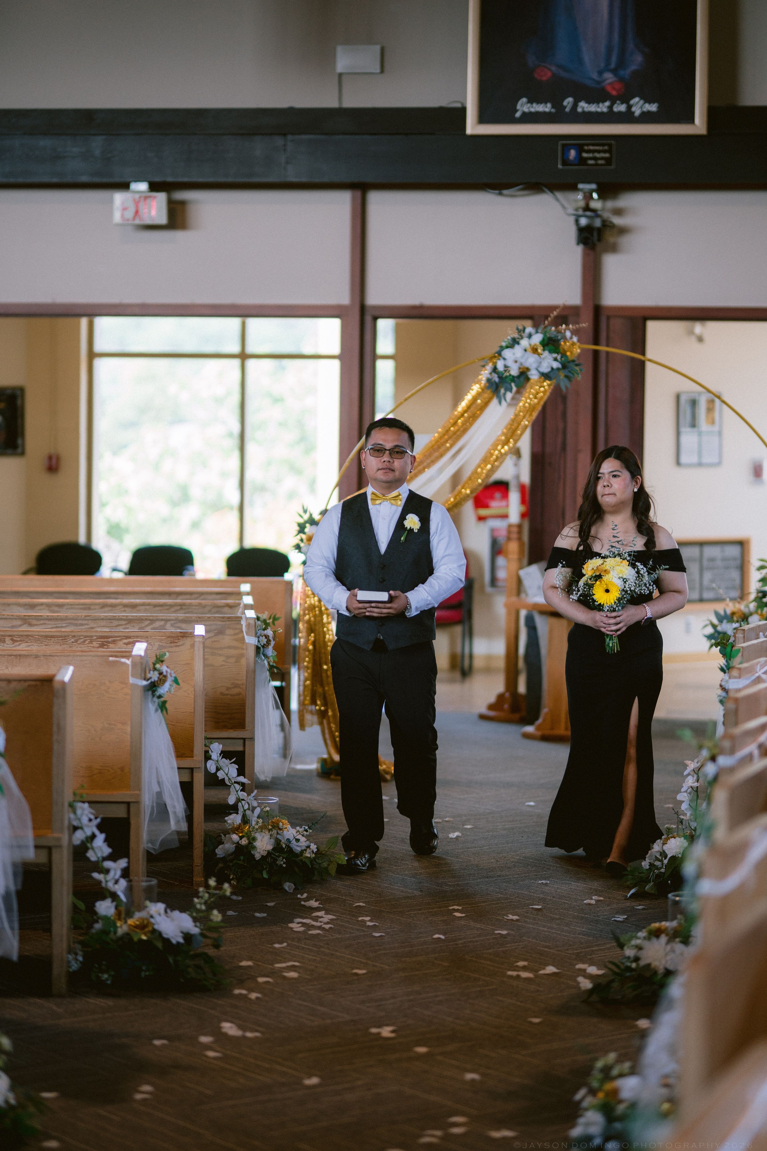 Wedding processional with groomsman and bridesmaid in black dress walking down church aisle with golden decor.