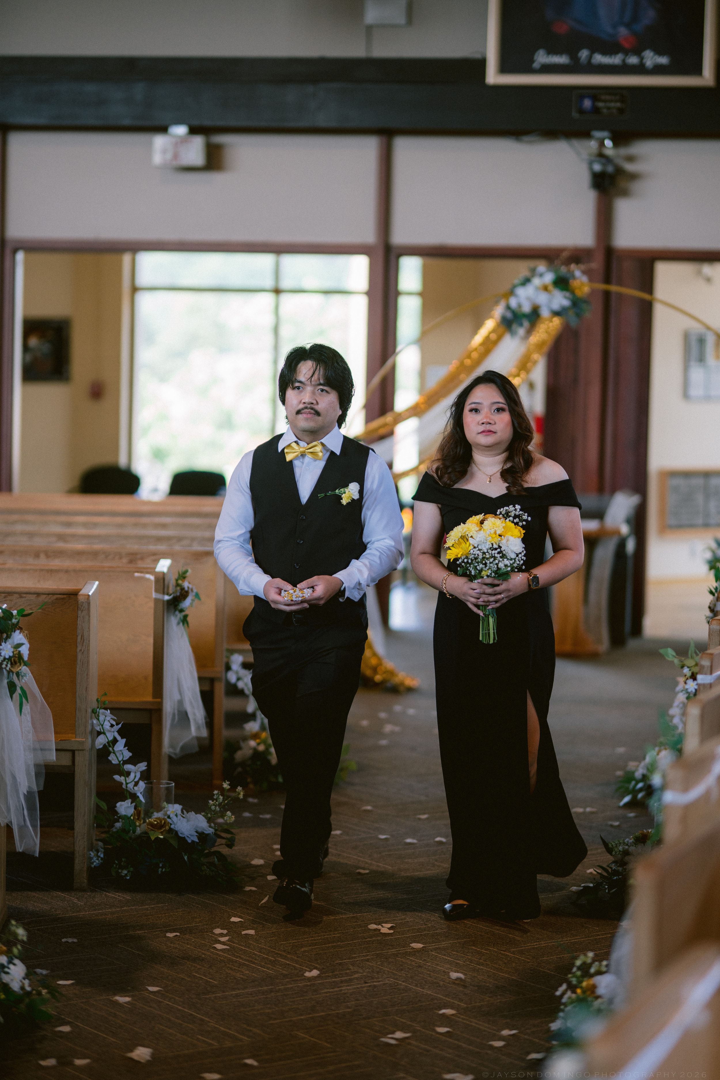 A groomsman and bridesmaid in black walk down a decorated church aisle during a wedding ceremony.