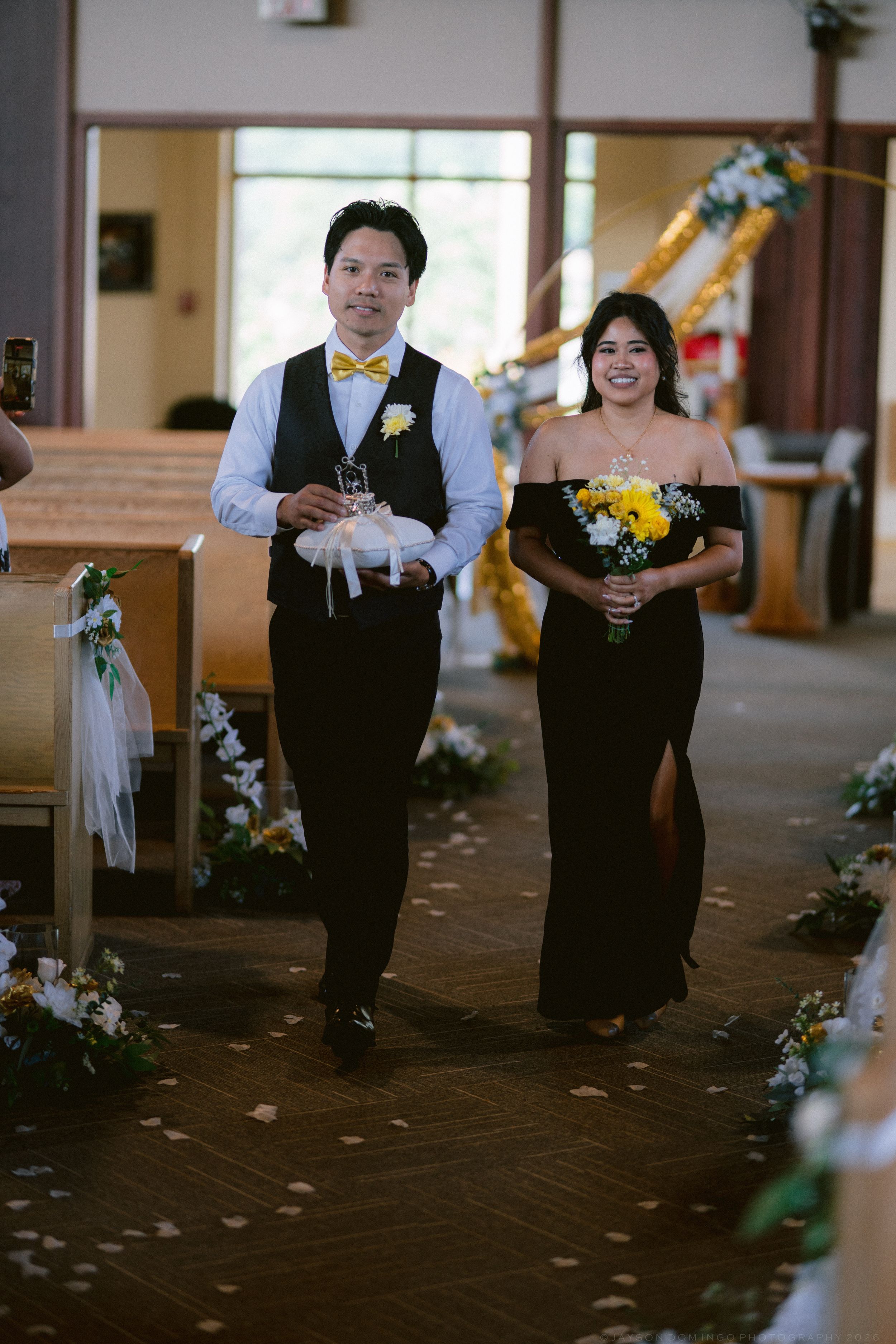 A groomsman and bridesmaid walk down the church aisle during a wedding ceremony with yellow floral decorations.