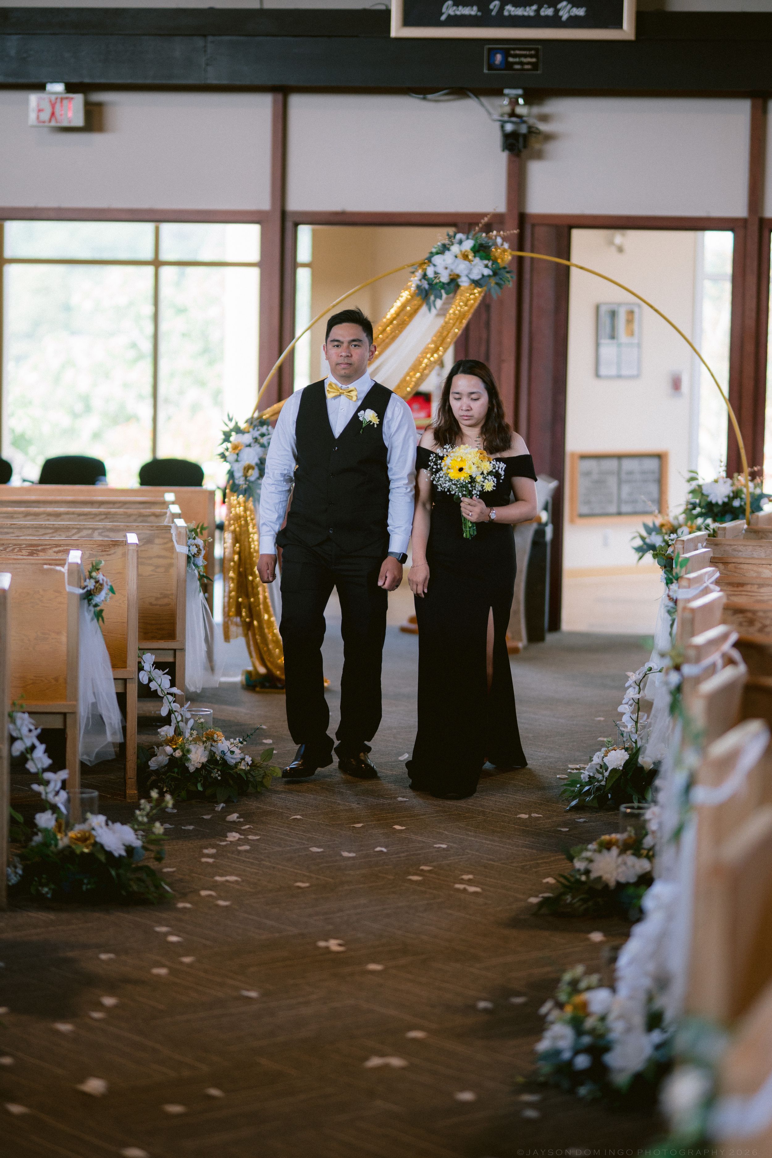 Groomsman and bridesmaid in black dress walk hand in hand down decorated church aisle at wedding.