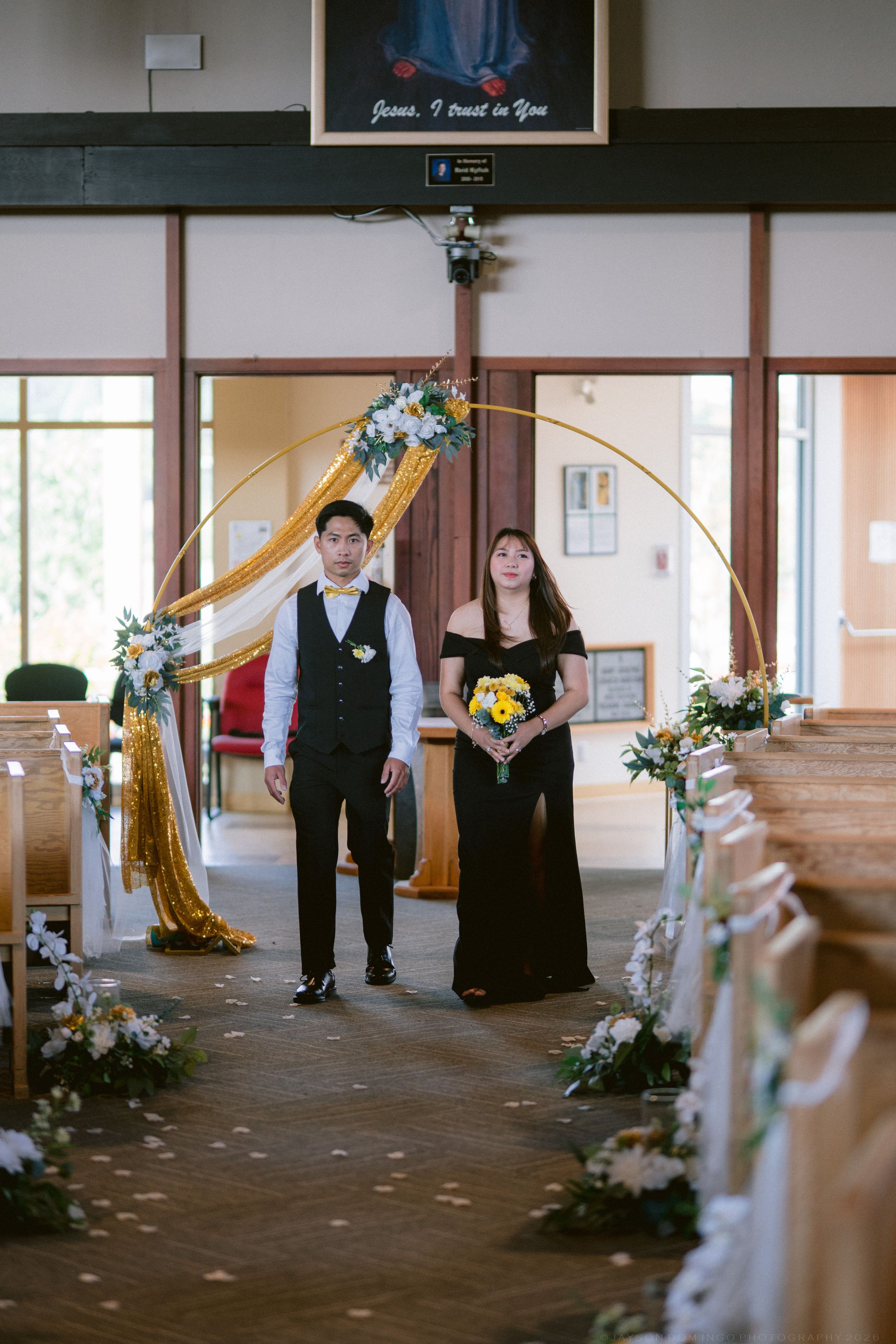 Groomsman and bridesmaid in black attire walking down a decorated church aisle with floral arrangements.