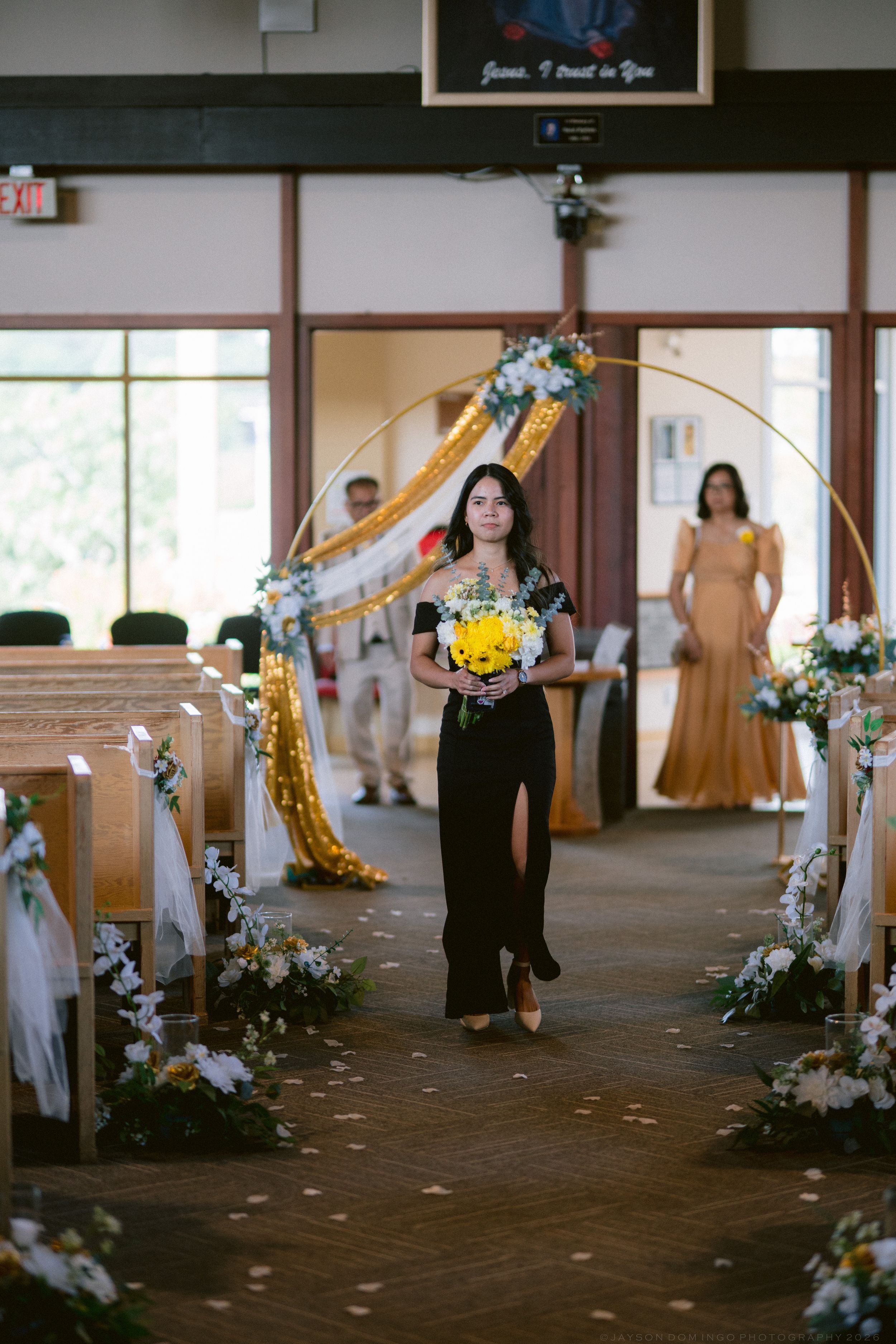 Bridesmaid in black dress walks down church aisle holding yellow bouquet during wedding ceremony.