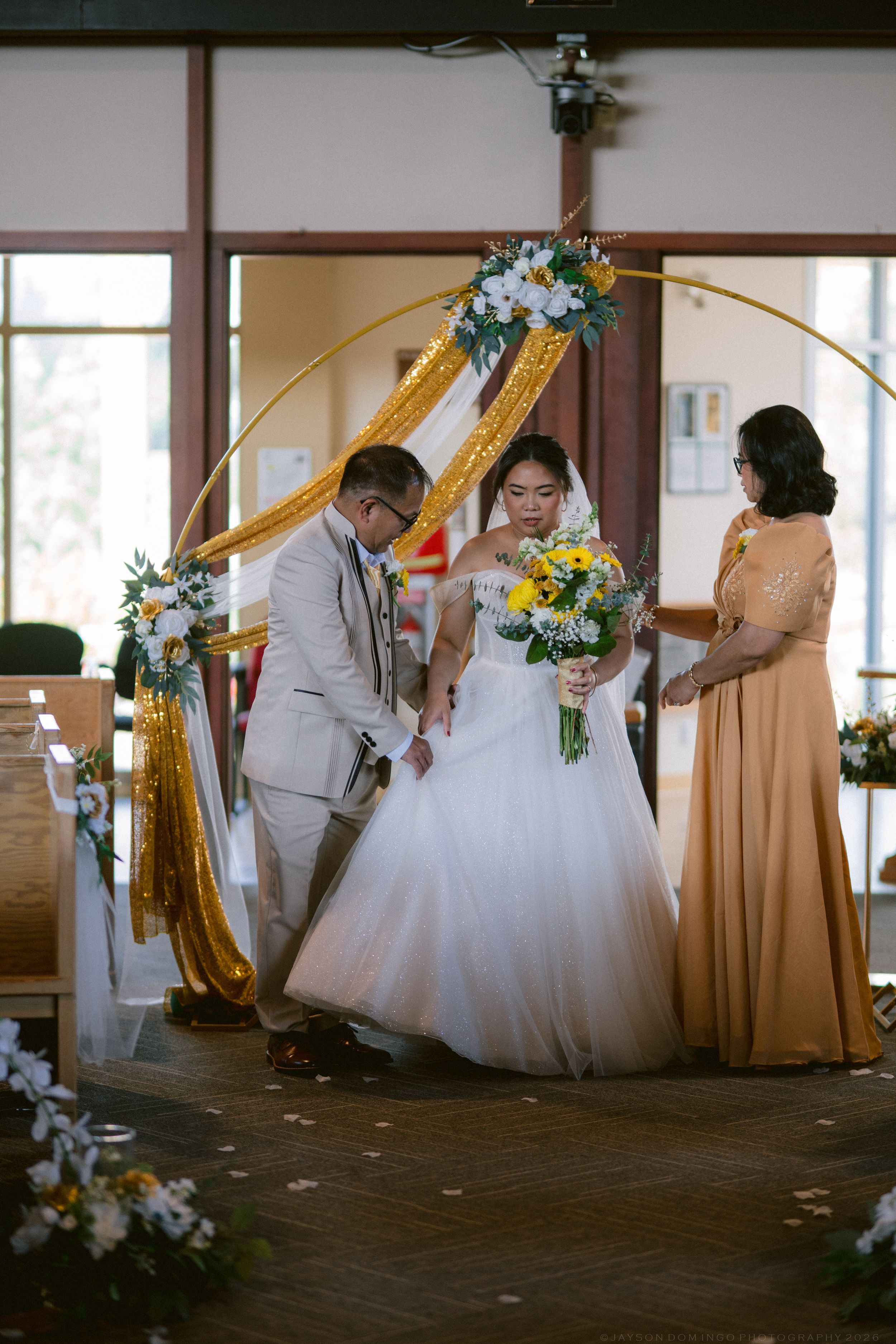 Bride in white ball gown being escorted to altar under golden floral arch by groom and family member.