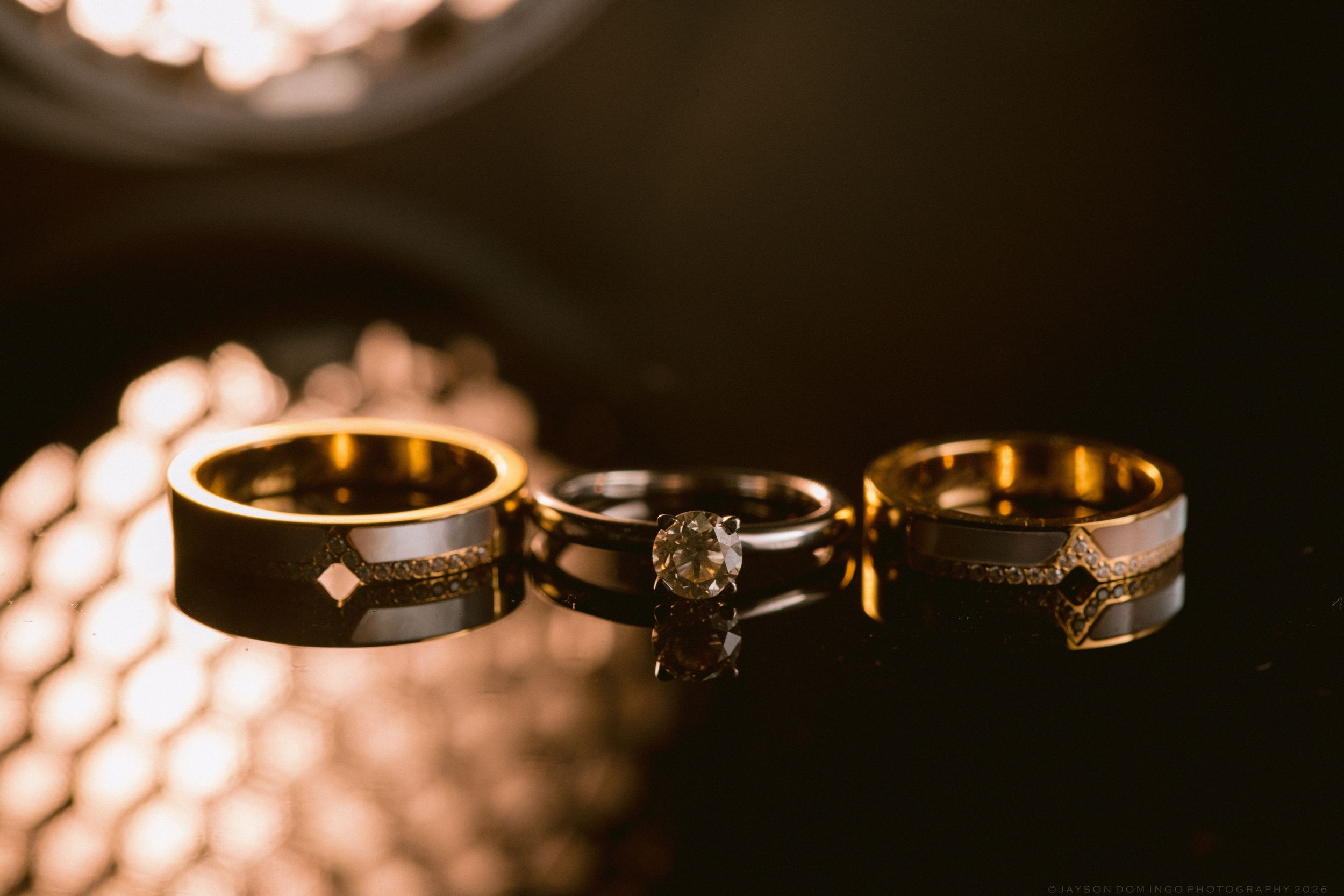 Three gold wedding rings and a diamond engagement ring displayed on a reflective black surface with warm bokeh lighting.