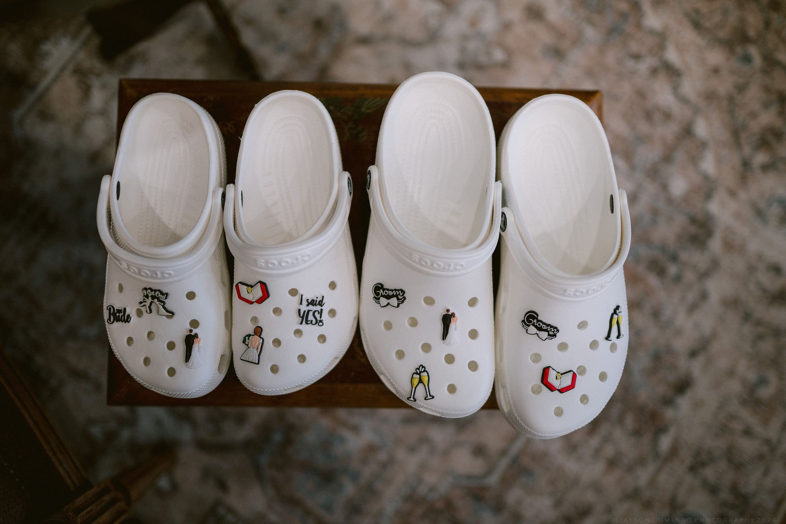 Four white Crocs with decorative jibbitz charms arranged on wooden tray for wedding party.