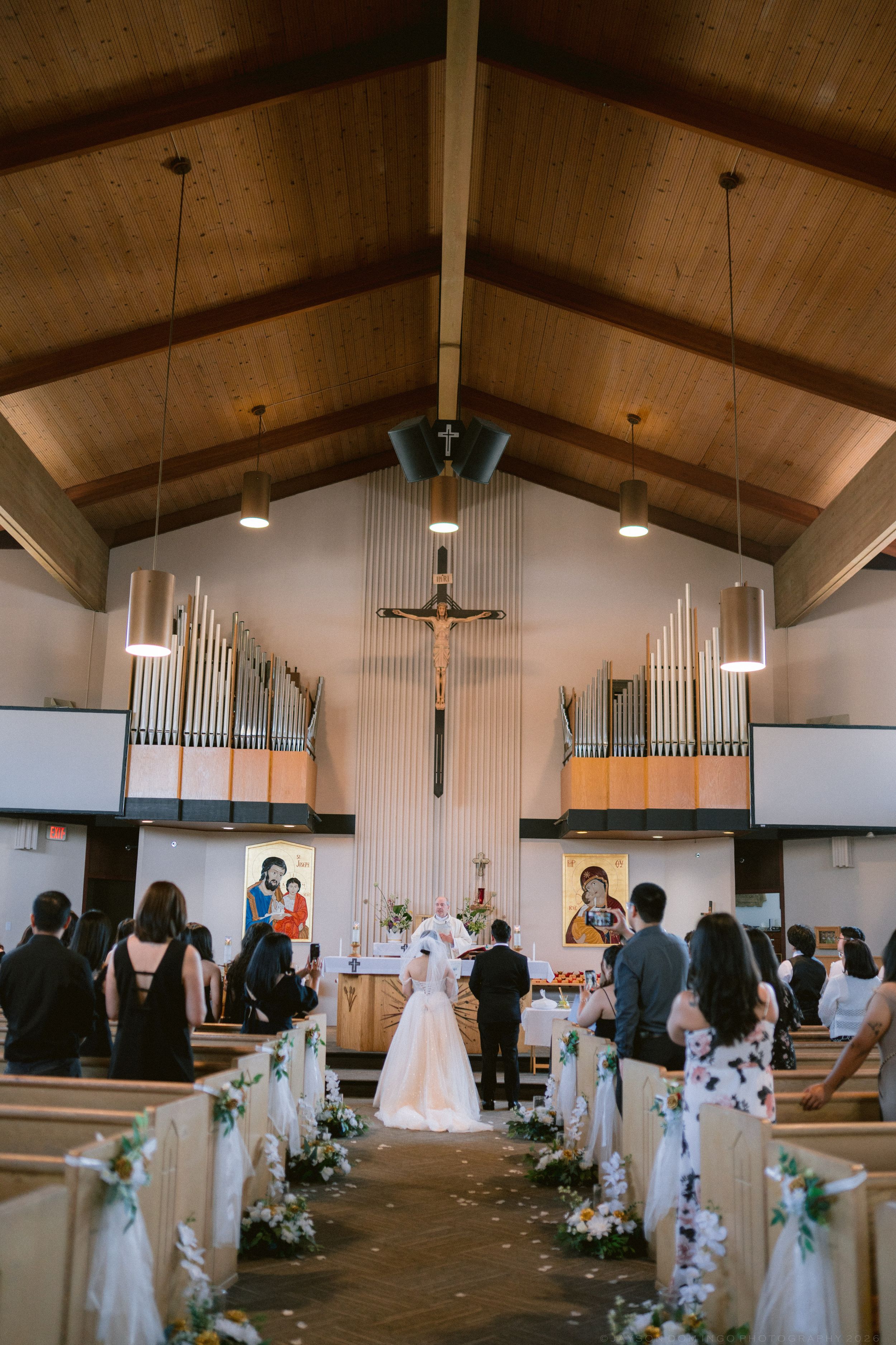 Wedding ceremony in a Catholic church with bride and groom at the altar, wooden ceiling, organ pipes, and floral-decorated pews.