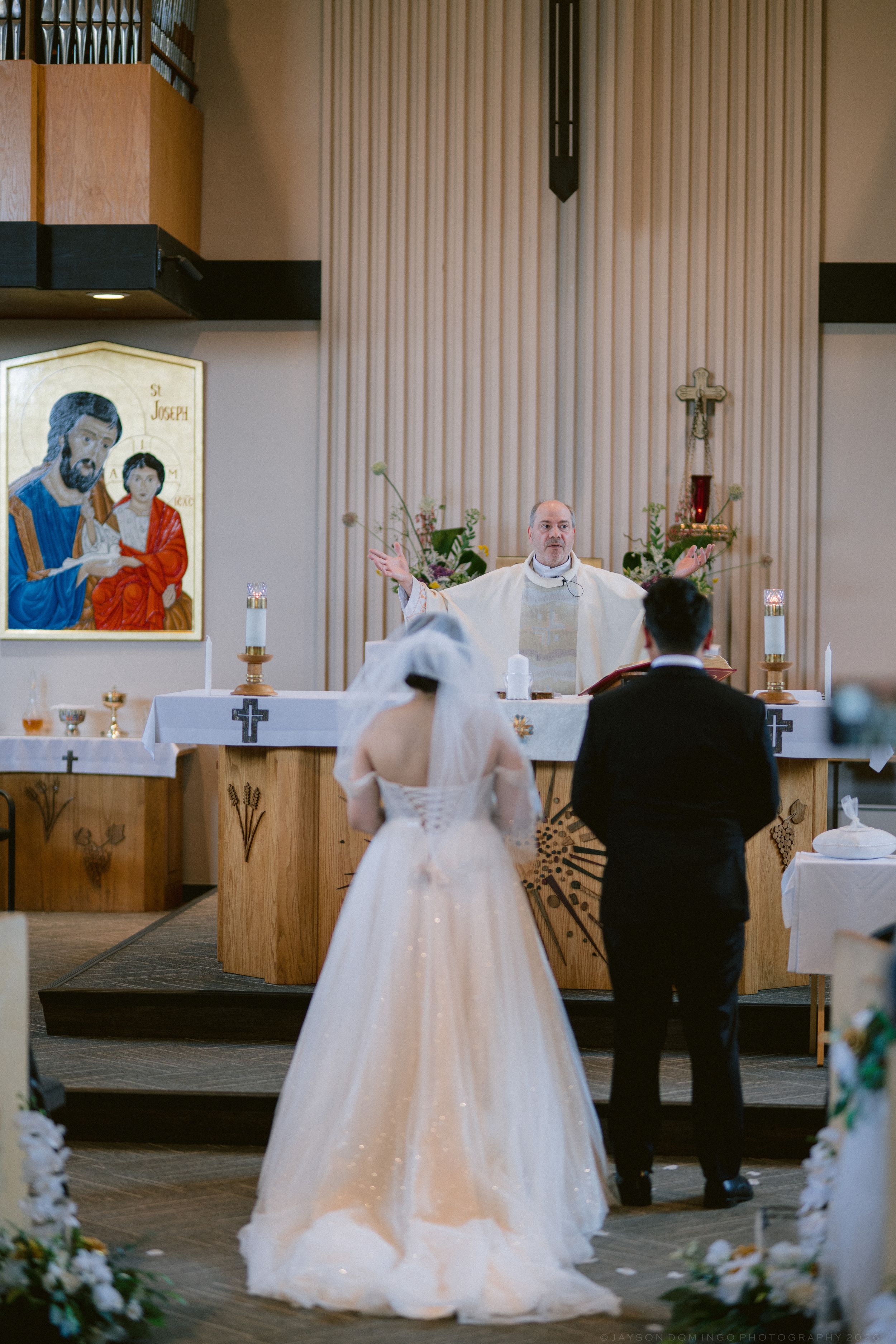 Bride and groom stand before priest at church altar during Catholic wedding ceremony with icon of Saint Joseph.