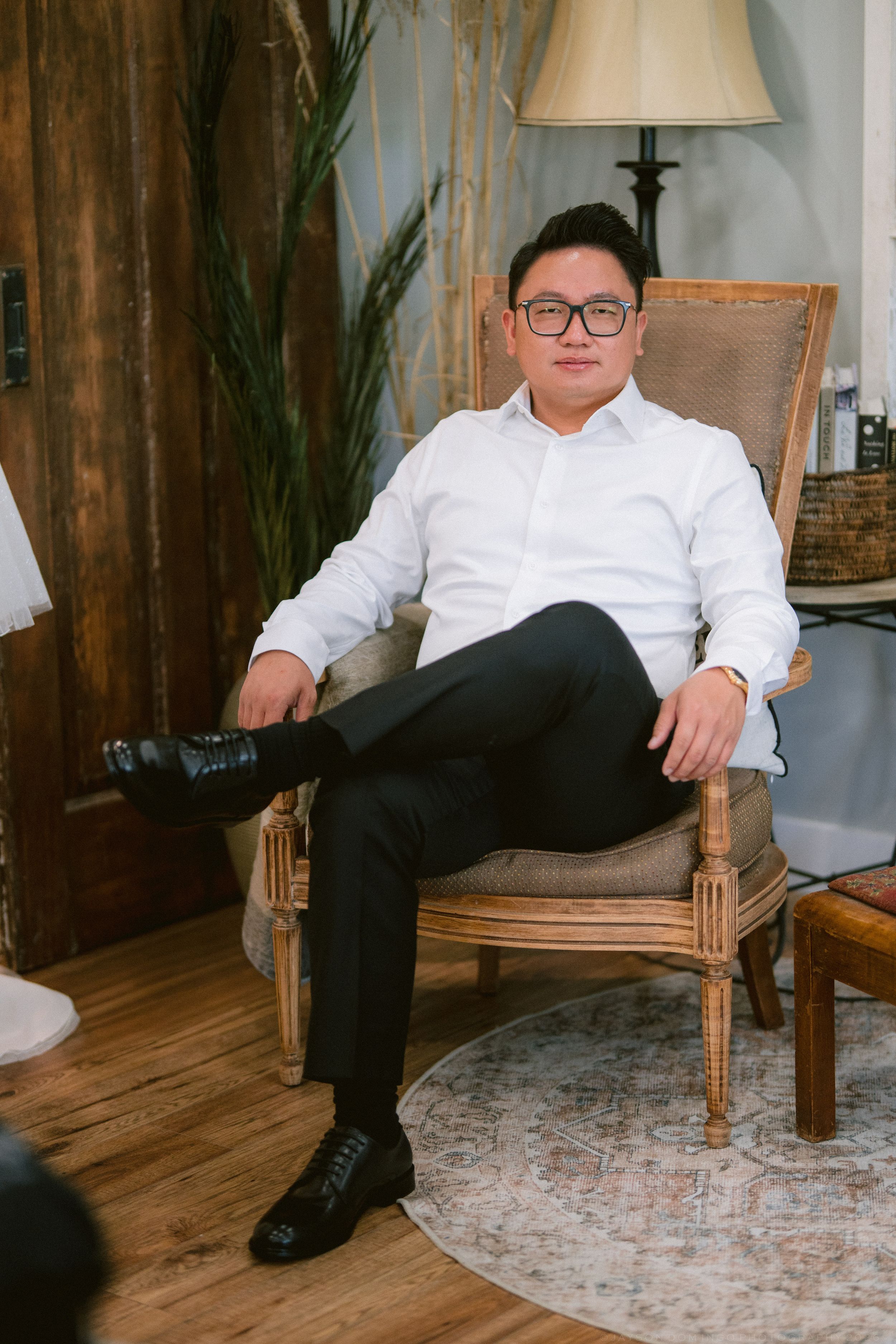Man in white shirt and black pants sits relaxed in wooden chair, crossing legs, wearing glasses in rustic interior.