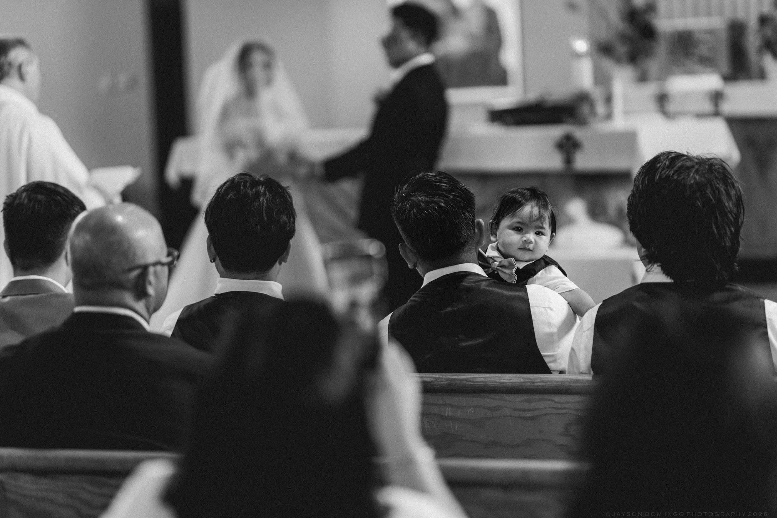 Black and white photo of wedding guests seated in church pews with small child looking toward the altar.
