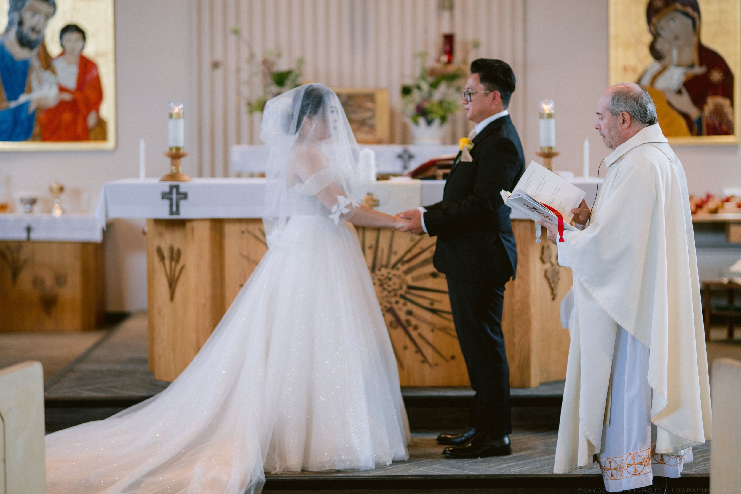 A bride and groom hold hands at the altar before a priest during a Catholic wedding ceremony.