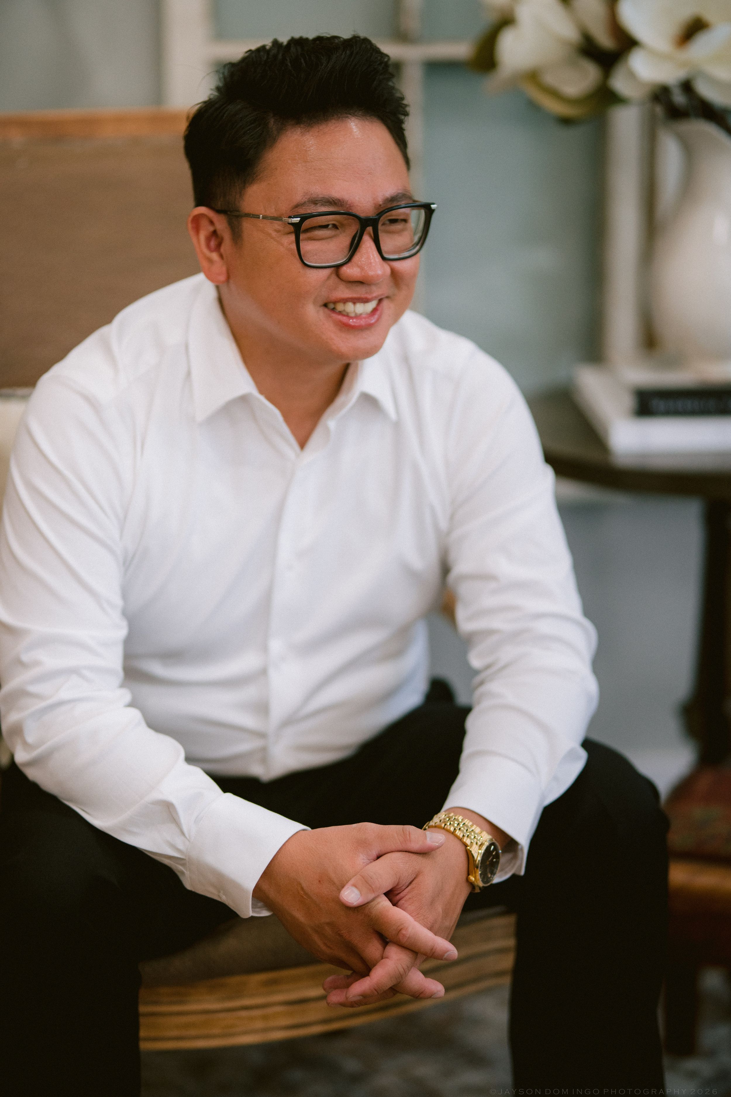 Smiling Asian man in white dress shirt and glasses, seated casually wearing a gold watch, in an elegant interior setting.
