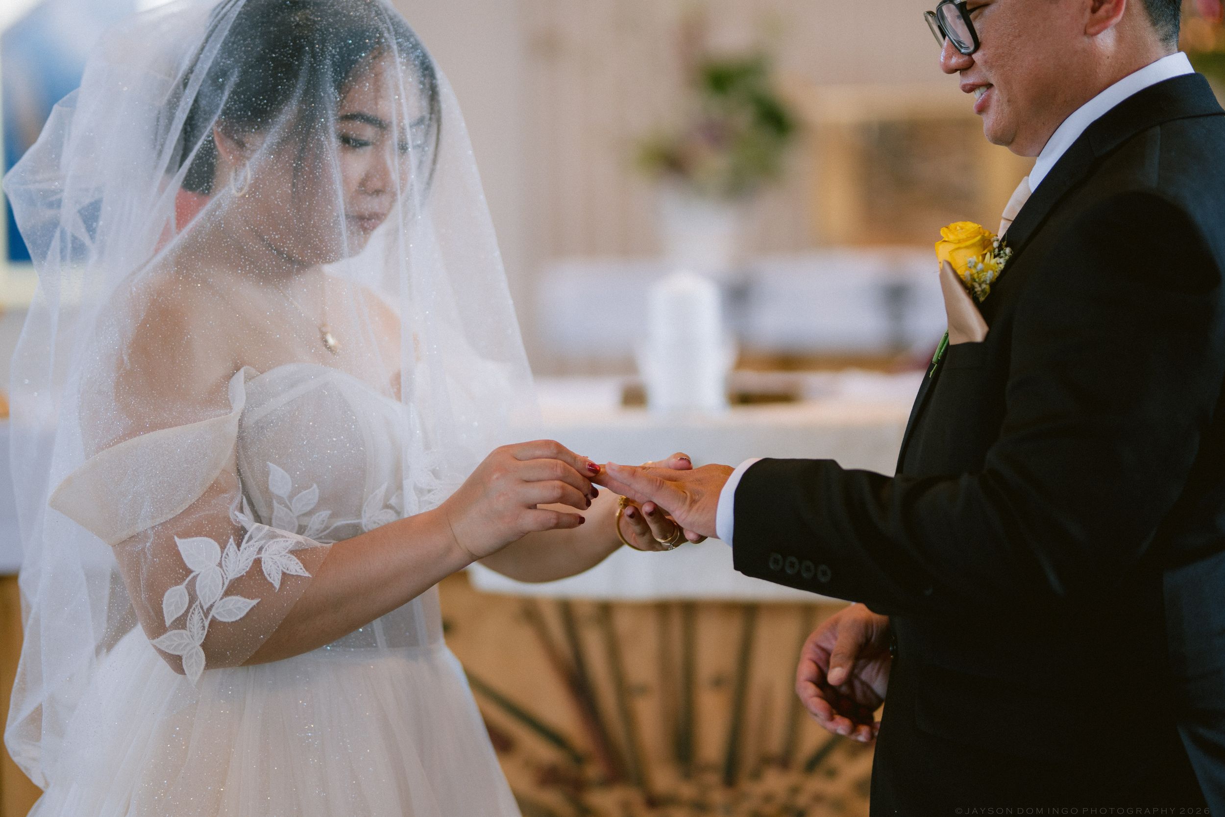 Bride in white lace gown and veil exchanges rings with groom in black tuxedo during wedding ceremony.
