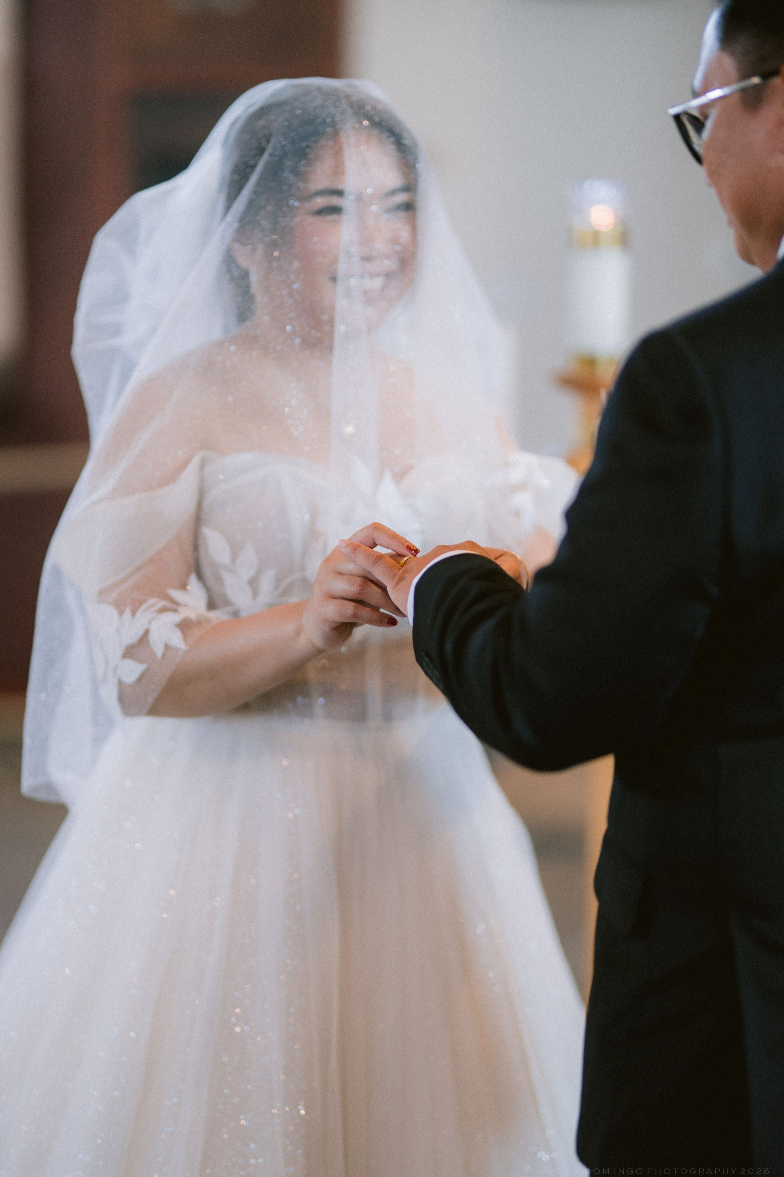 Smiling bride in a sparkly ballgown and veil exchanges rings with groom during church ceremony.