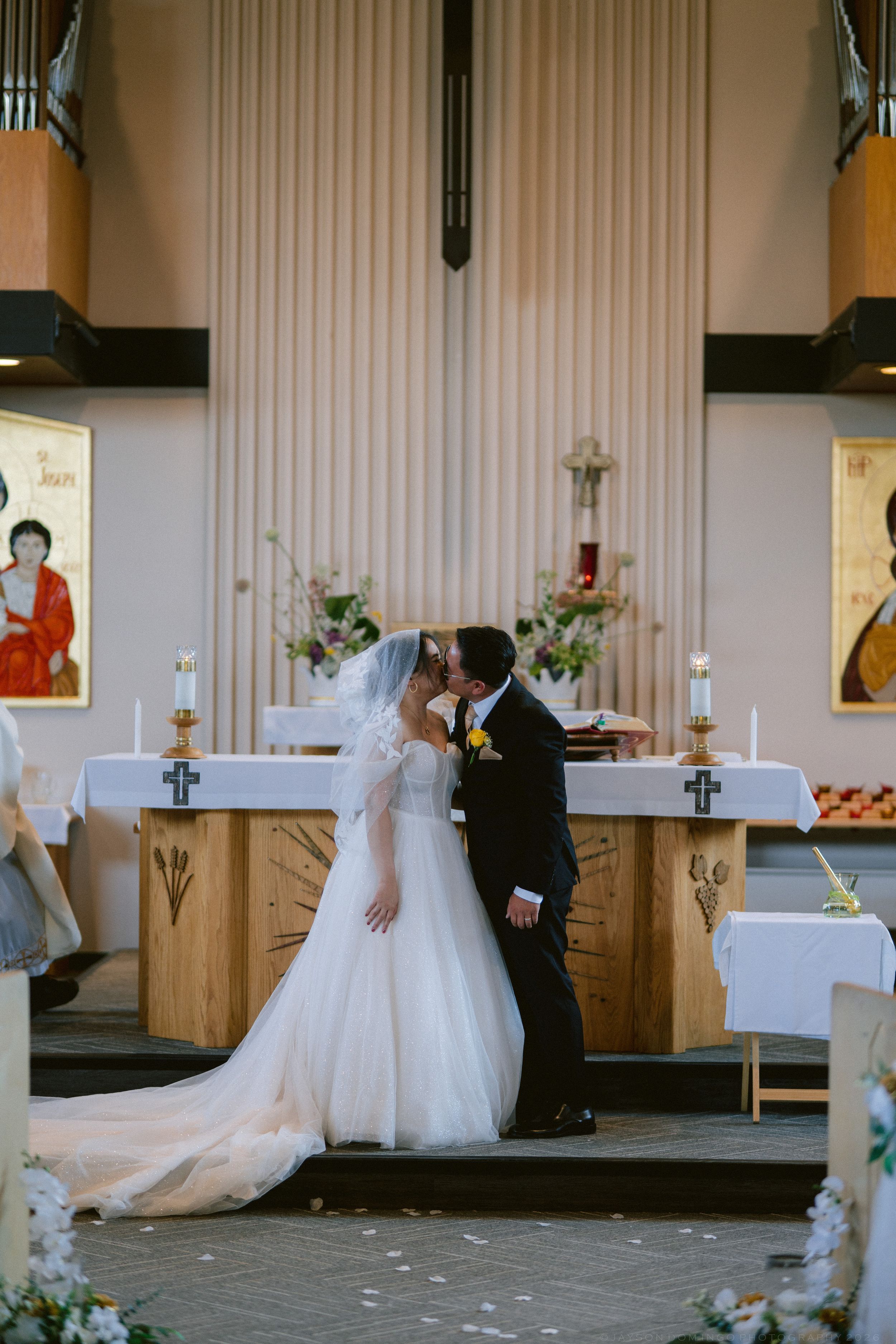 Bride and groom share first kiss at church altar during wedding ceremony with floral arrangements and religious icons.