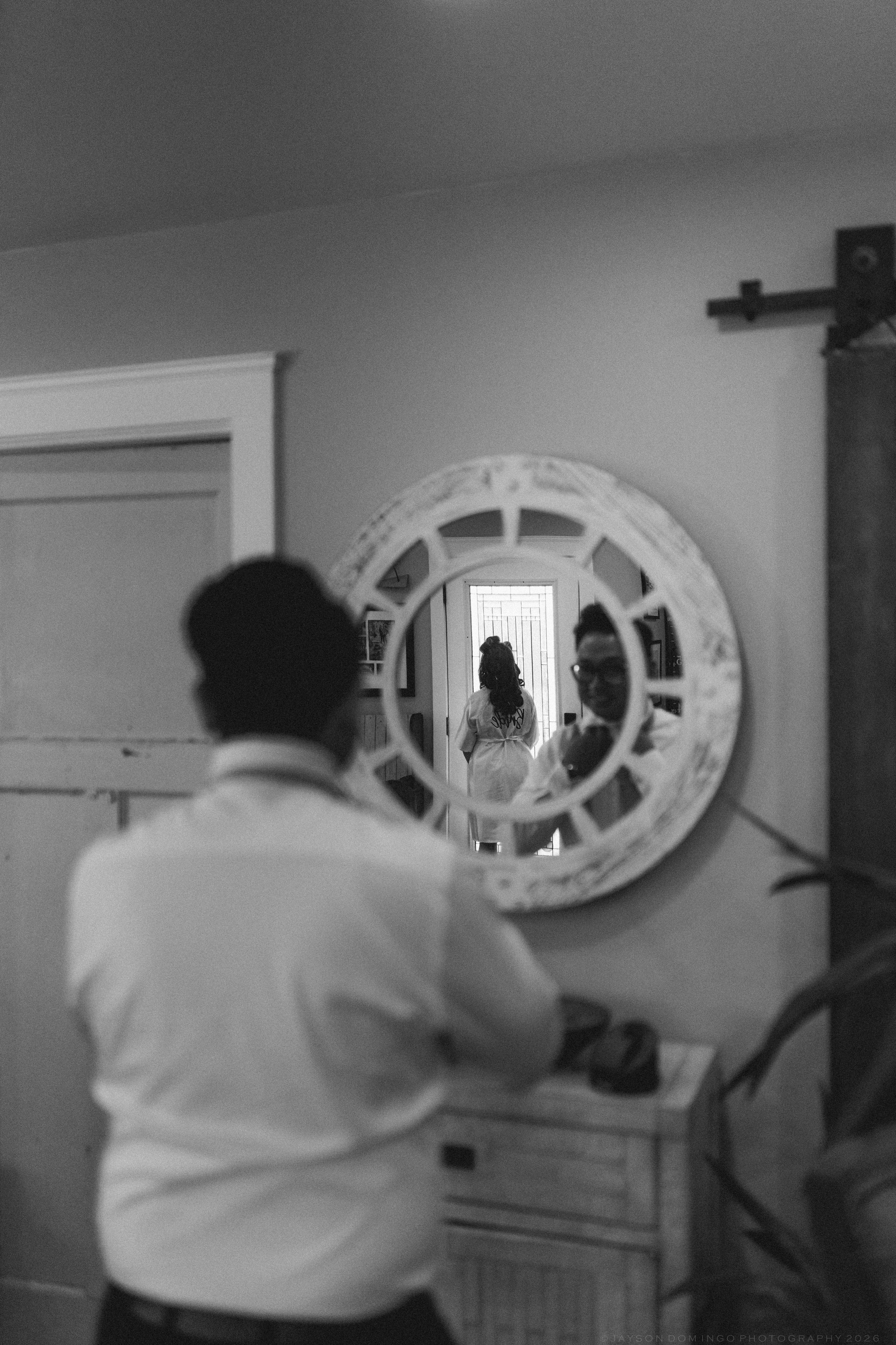 Groom adjusting his tie while viewing his reflection in a decorative round mirror in black and white.