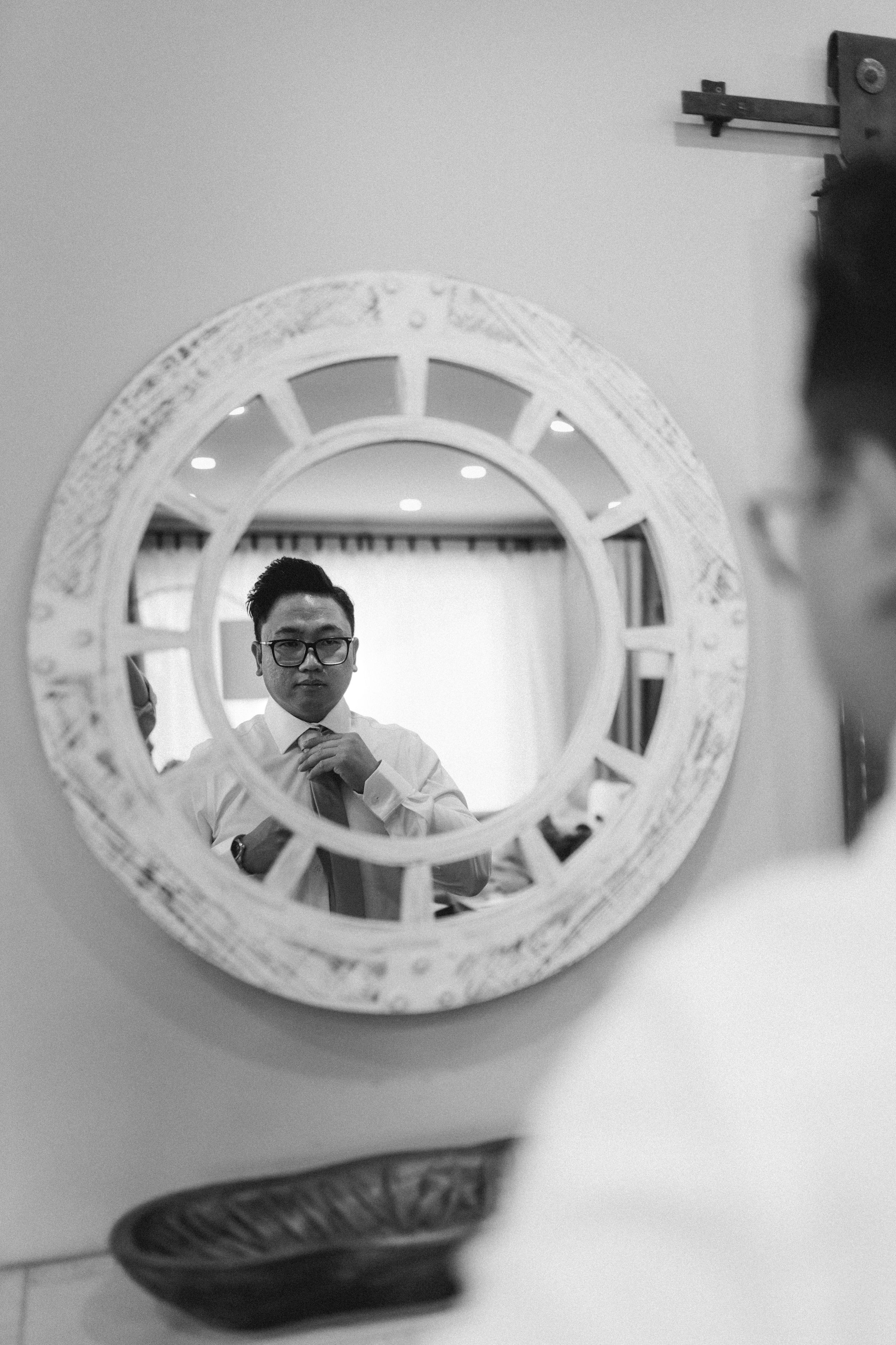 Groom adjusting tie reflected in ornate round mirror, black and white wedding preparation photo.