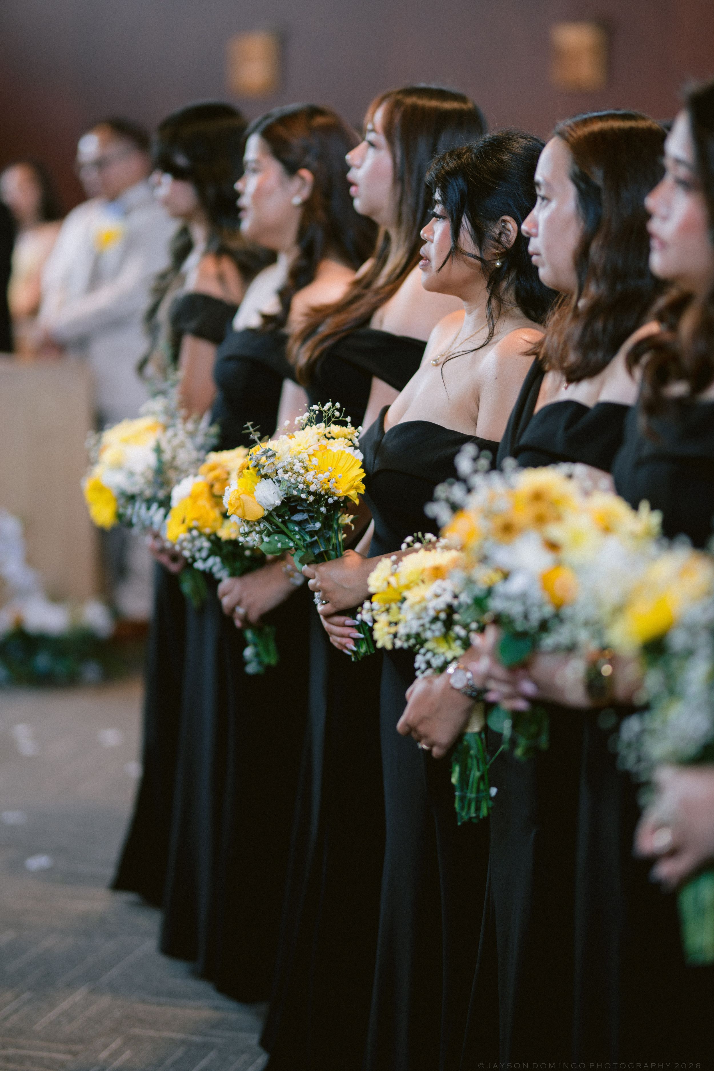 Bridesmaids in black off-shoulder gowns holding vibrant yellow flower bouquets during wedding ceremony.