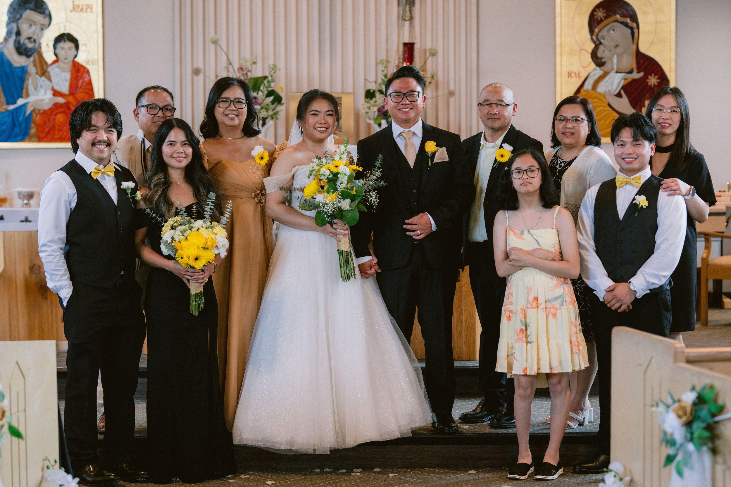 Wedding party group photo inside church featuring bride in white gown and groom with yellow floral accents.