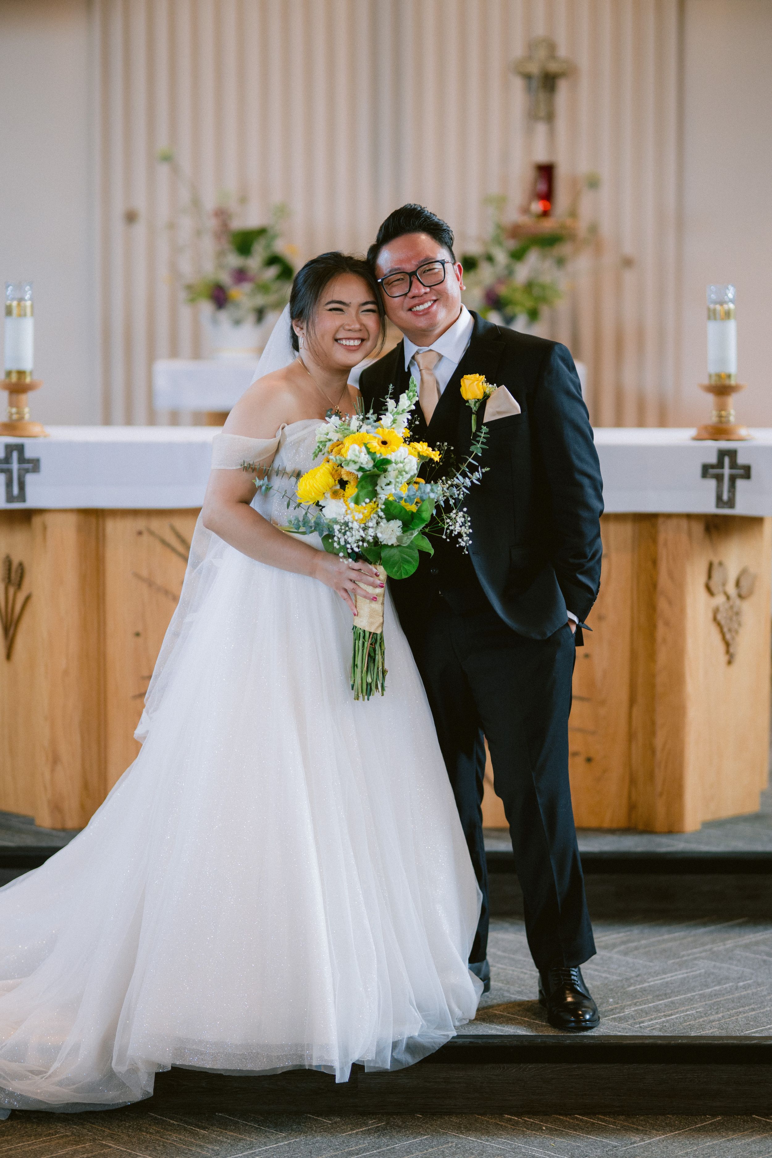 Smiling bride in off-shoulder gown and groom in black suit pose together at church altar with yellow bouquet.