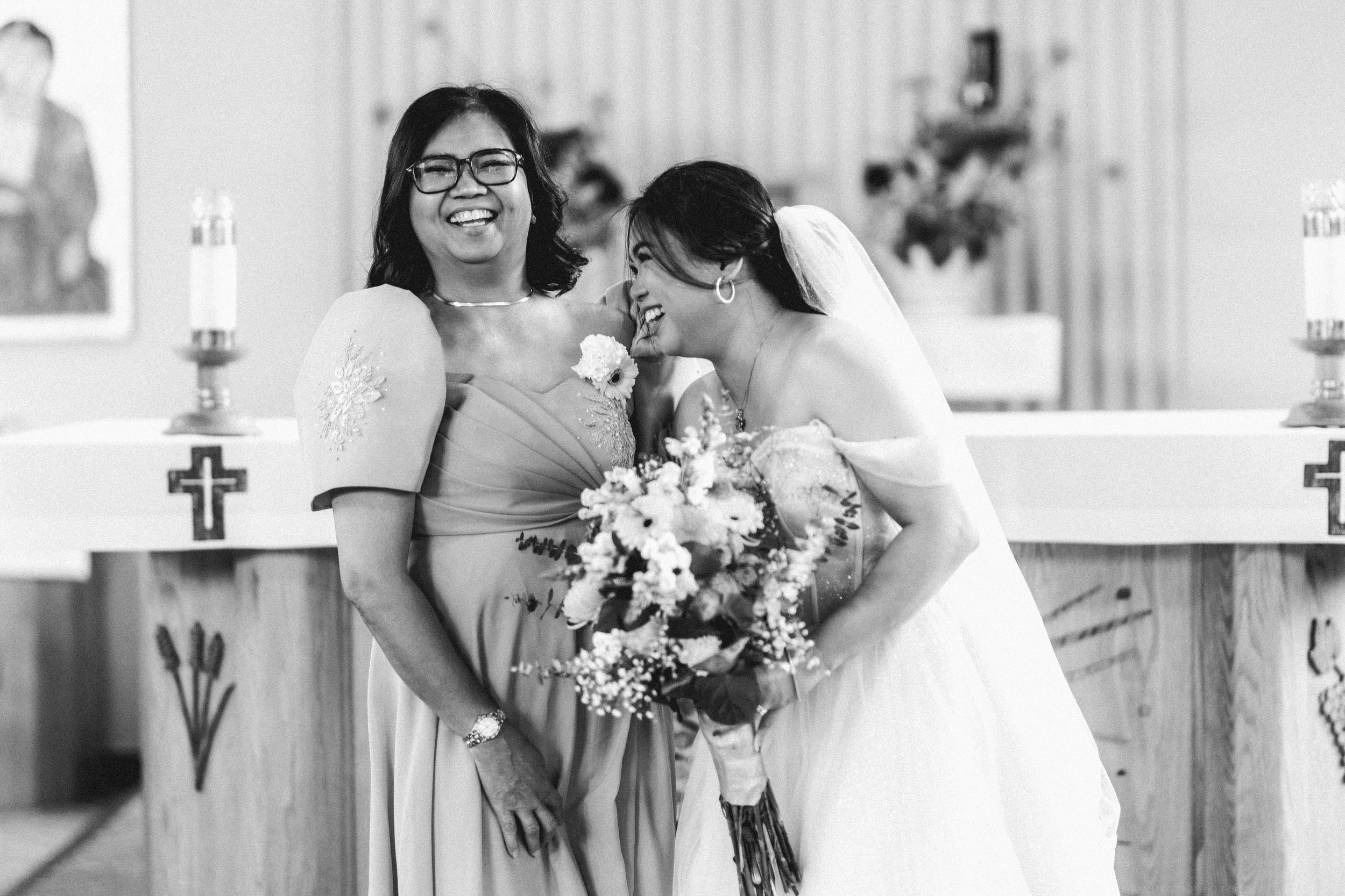 Bride in white gown laughs with smiling bridesmaid in champagne dress at church altar, black and white.