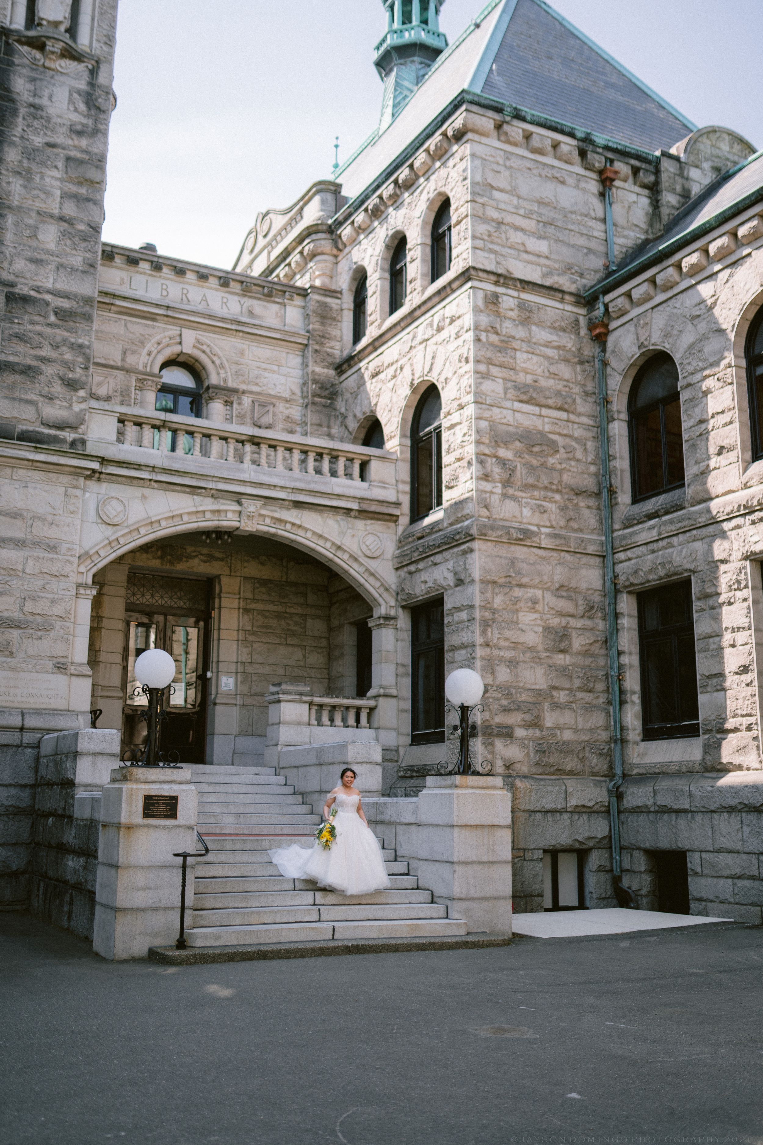 Bride in white ball gown descends stone steps of a grand historic building holding a yellow bouquet.