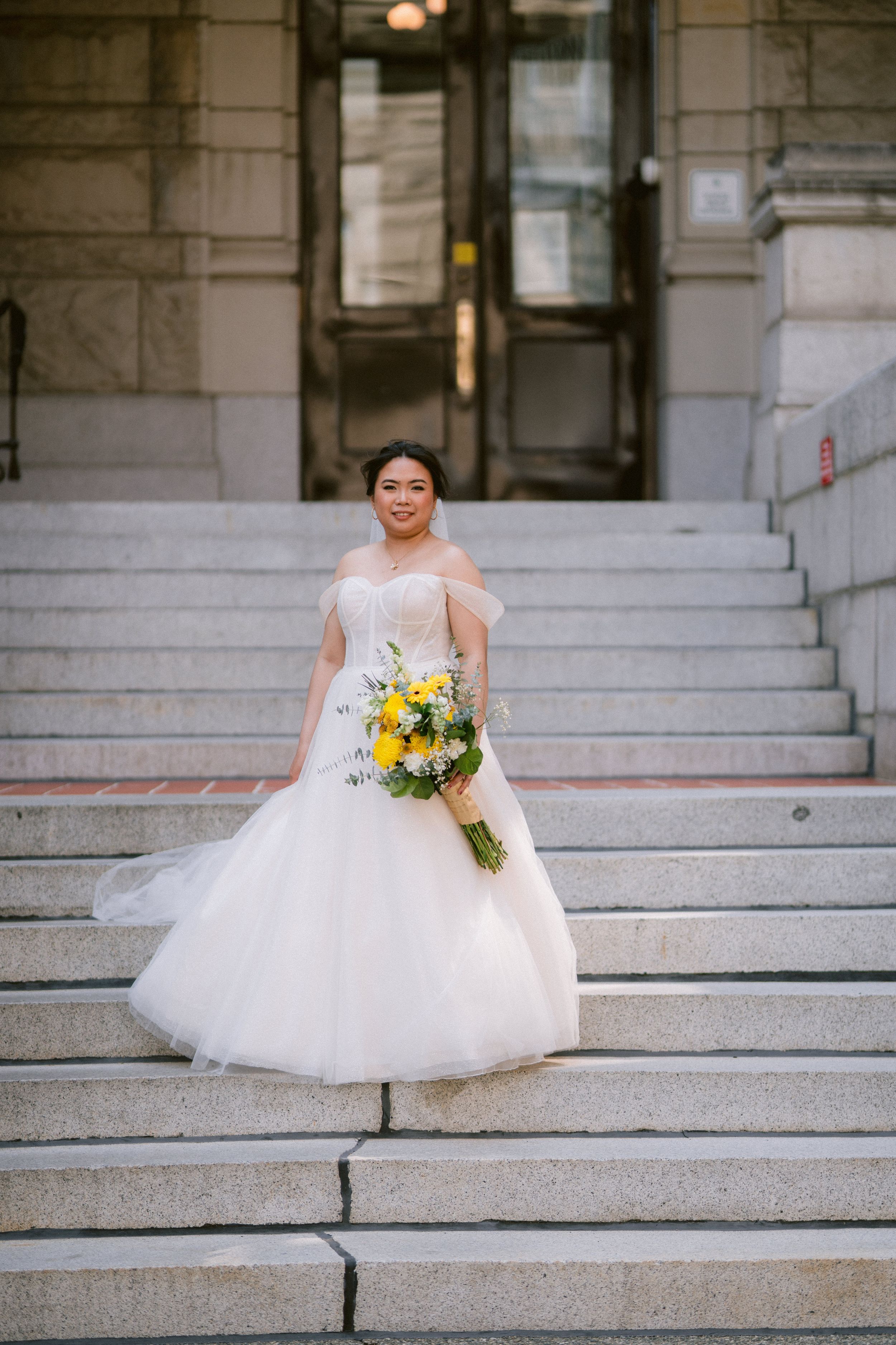 Bride in strapless ball gown holding yellow bouquet poses on stone steps of elegant building.