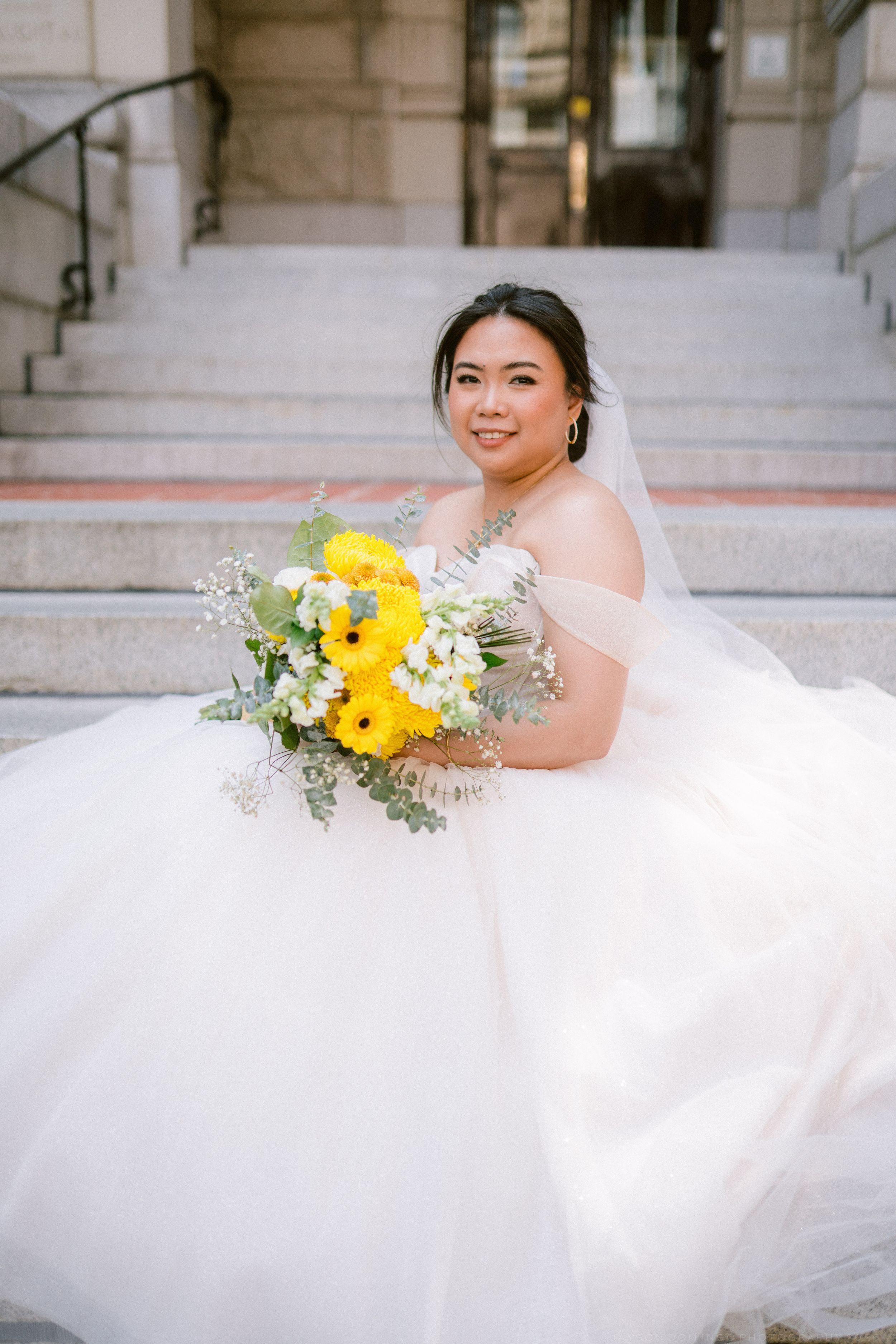Smiling Asian bride in off-shoulder white ballgown holds yellow sunflower bouquet on stone steps.