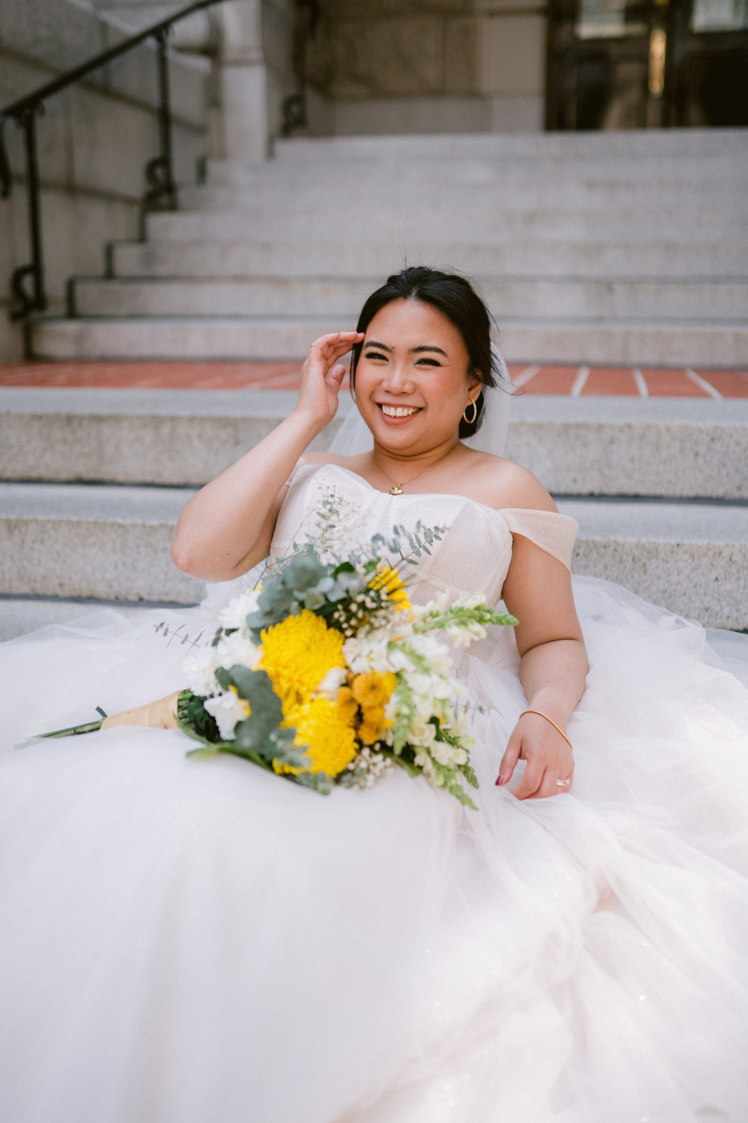 Radiant Asian bride in white tulle ballgown sits on stone steps with yellow and white floral bouquet.