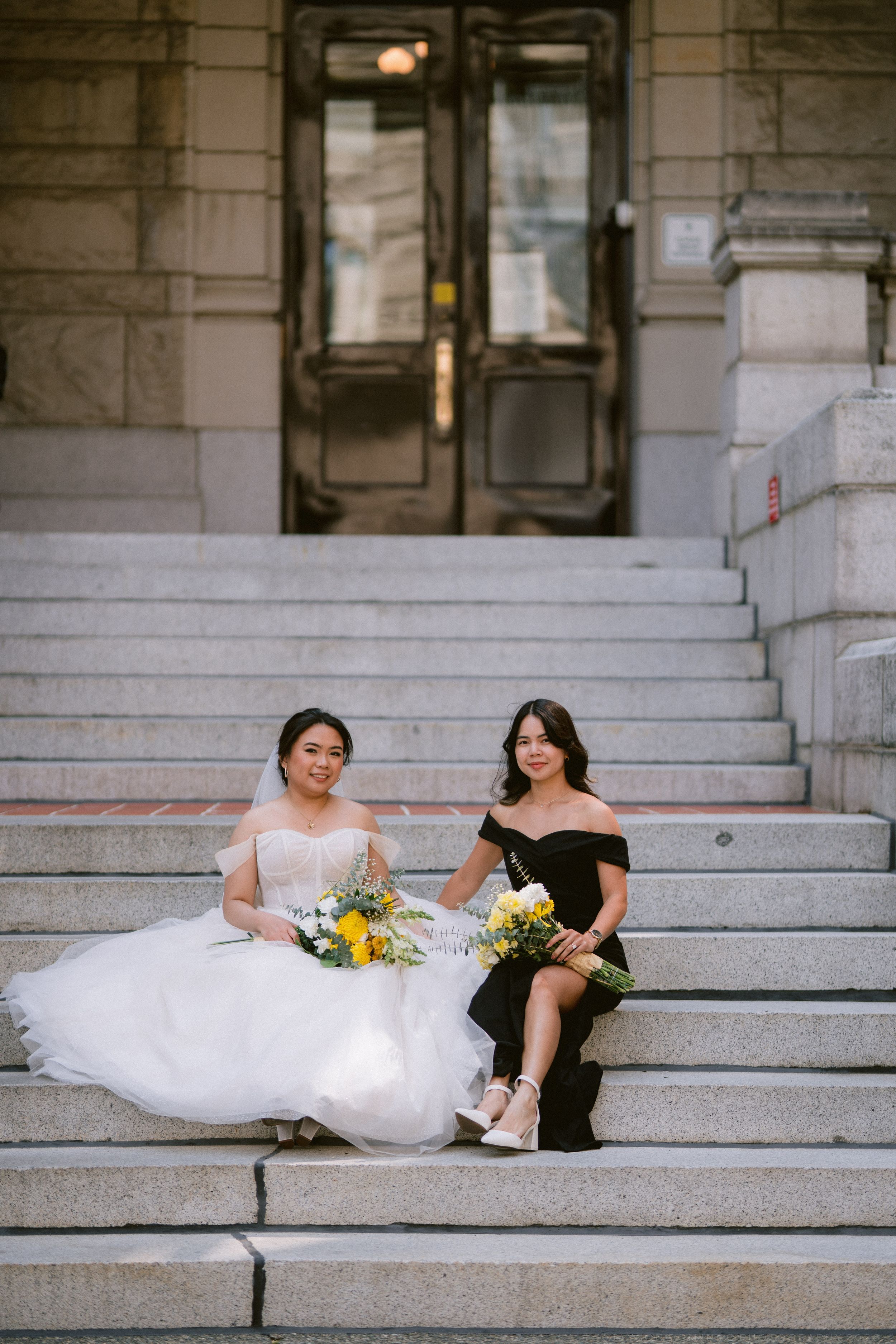 Bride in white gown and bridesmaid in black dress sitting on stone steps holding yellow floral bouquets.