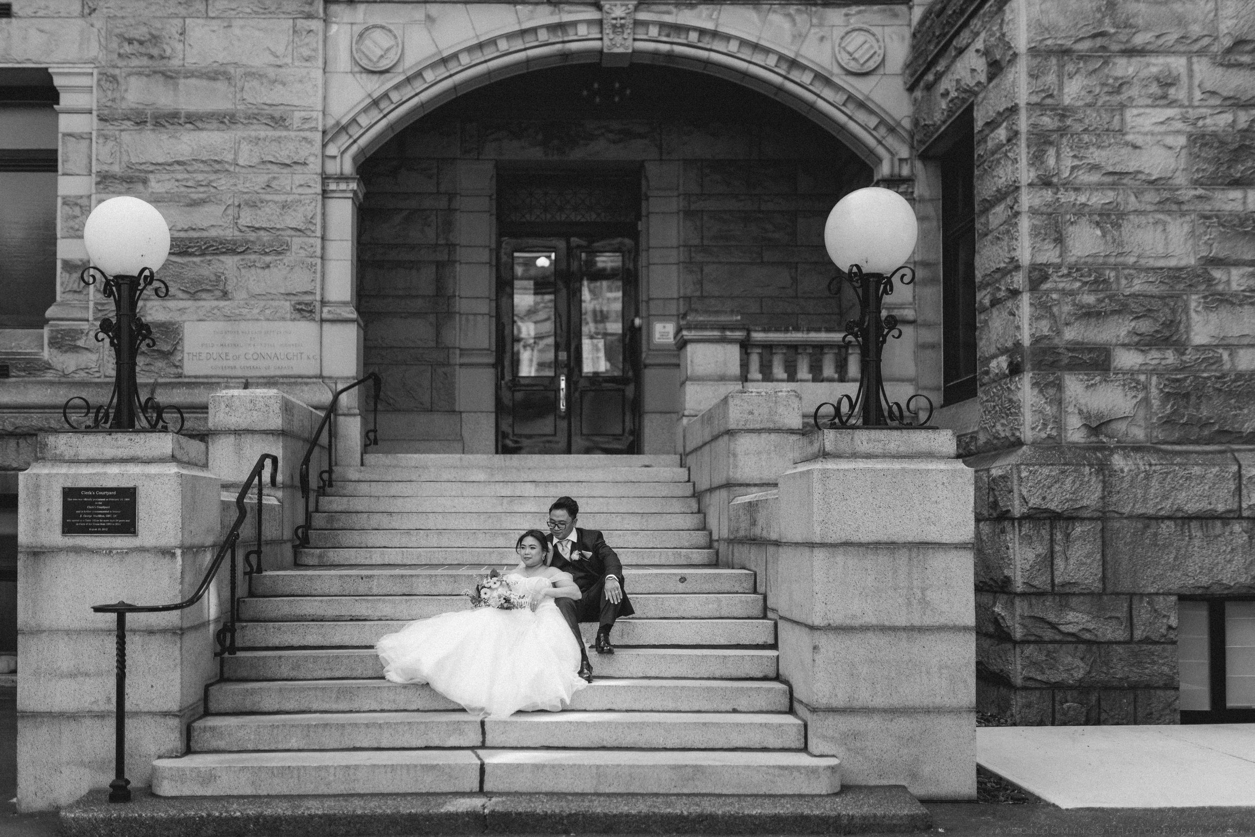 Black and white photo of bride and groom sitting on steps of grand stone building after wedding ceremony.