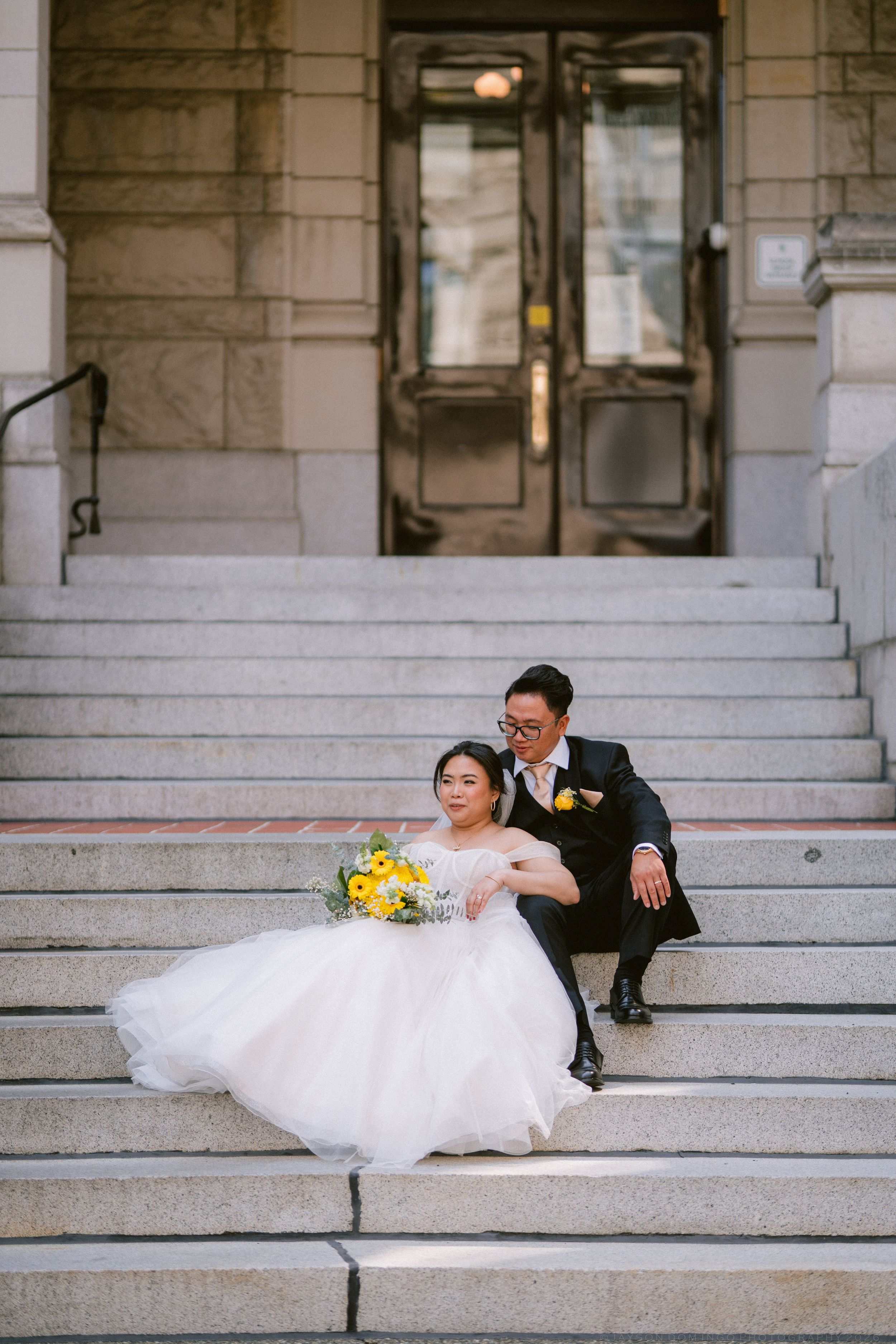 Bride and groom sitting on stone steps of historic building, bride holding yellow bouquet.