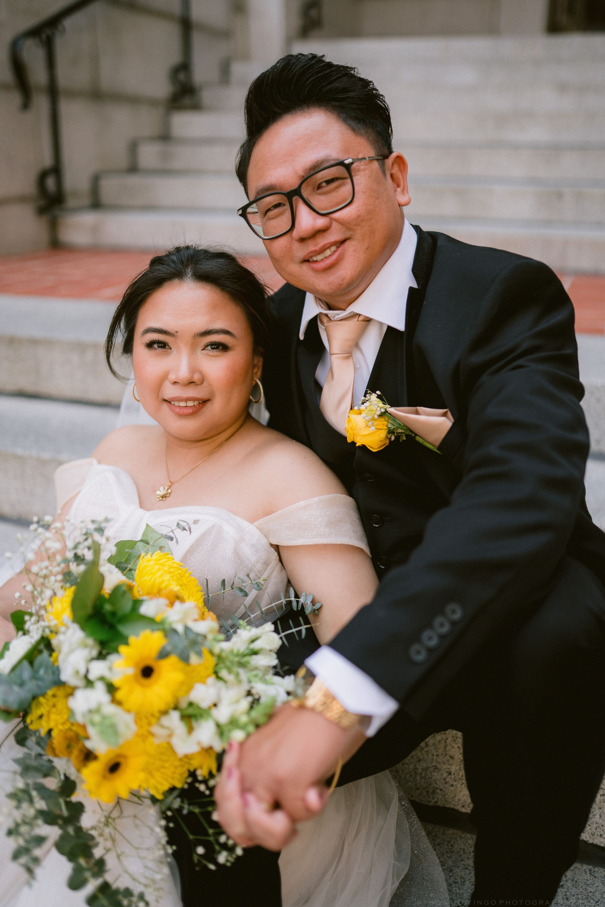 Smiling Asian bride and groom sitting on stone steps, bride holding yellow sunflower bouquet, groom in black suit with peach tie.