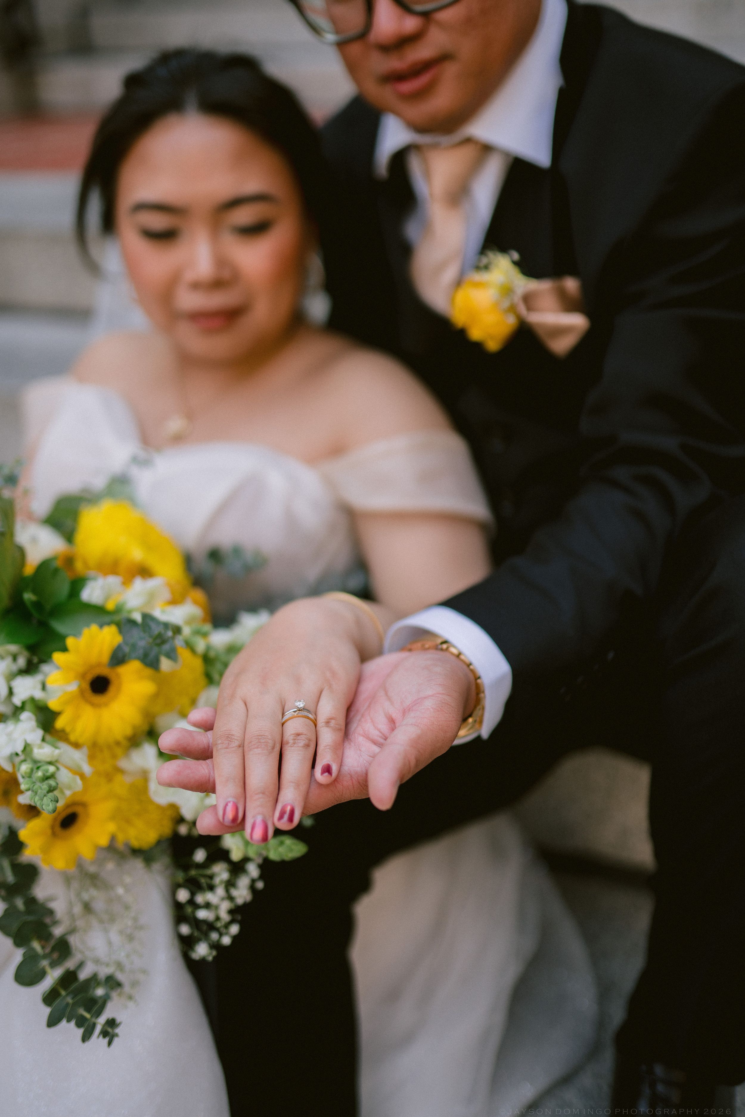 Newlyweds display matching gold wedding rings, bride holding yellow sunflower bouquet in white off-shoulder gown.