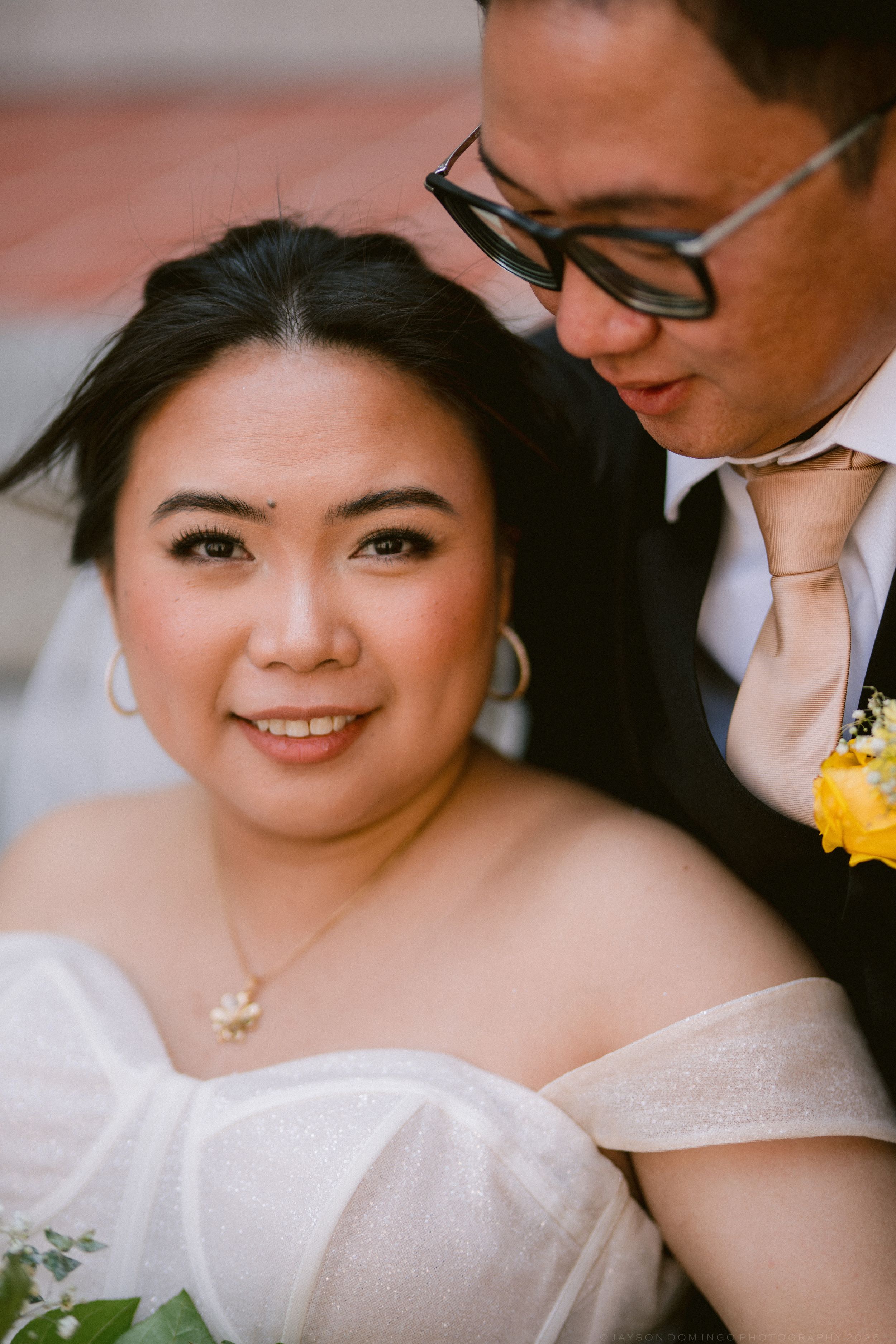 Close-up portrait of smiling Asian bride in off-shoulder gown with groom in glasses nuzzling her cheek.