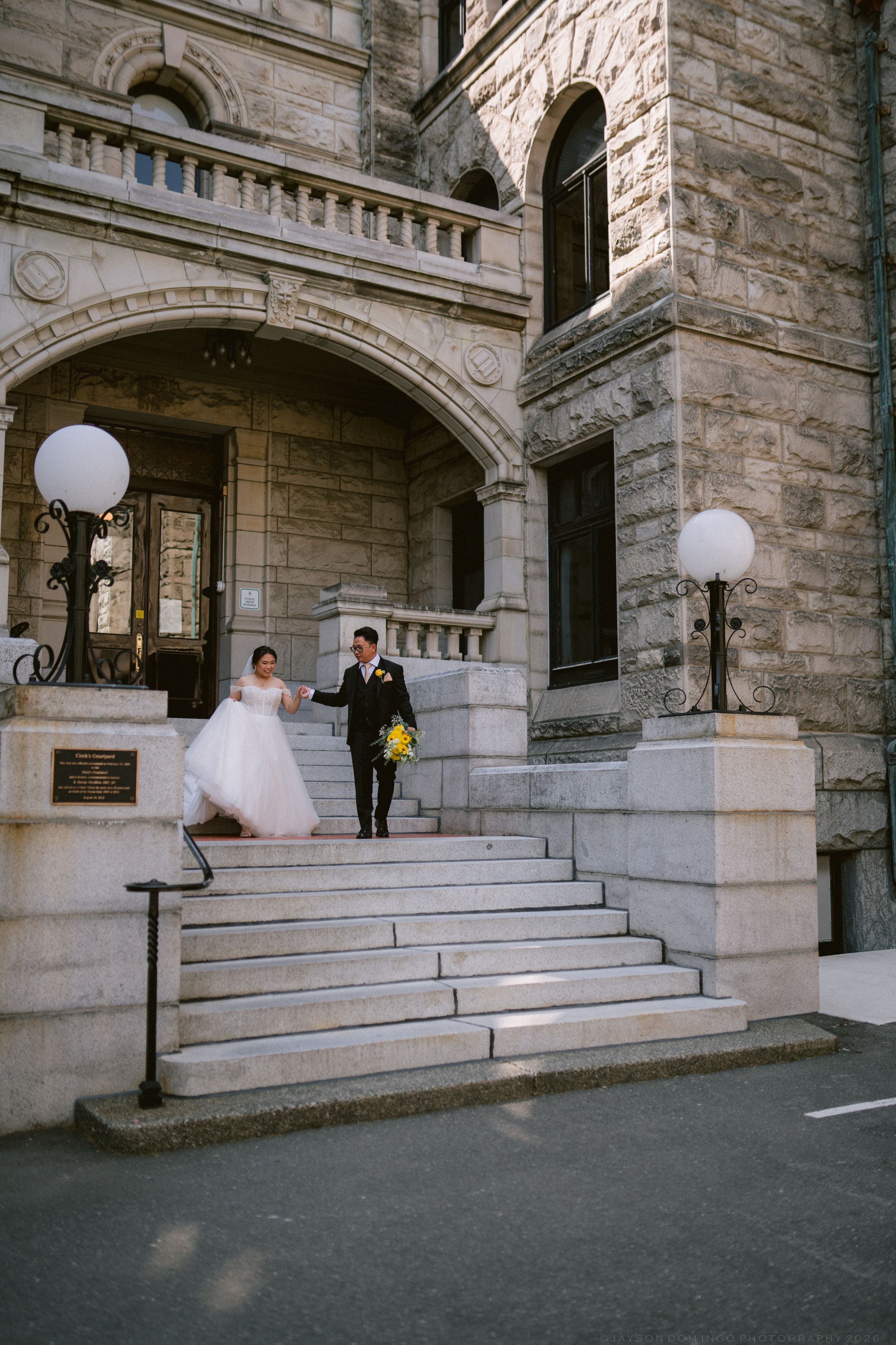 Bride in flowing pink gown and groom with yellow bouquet descend stone steps of historic building together.