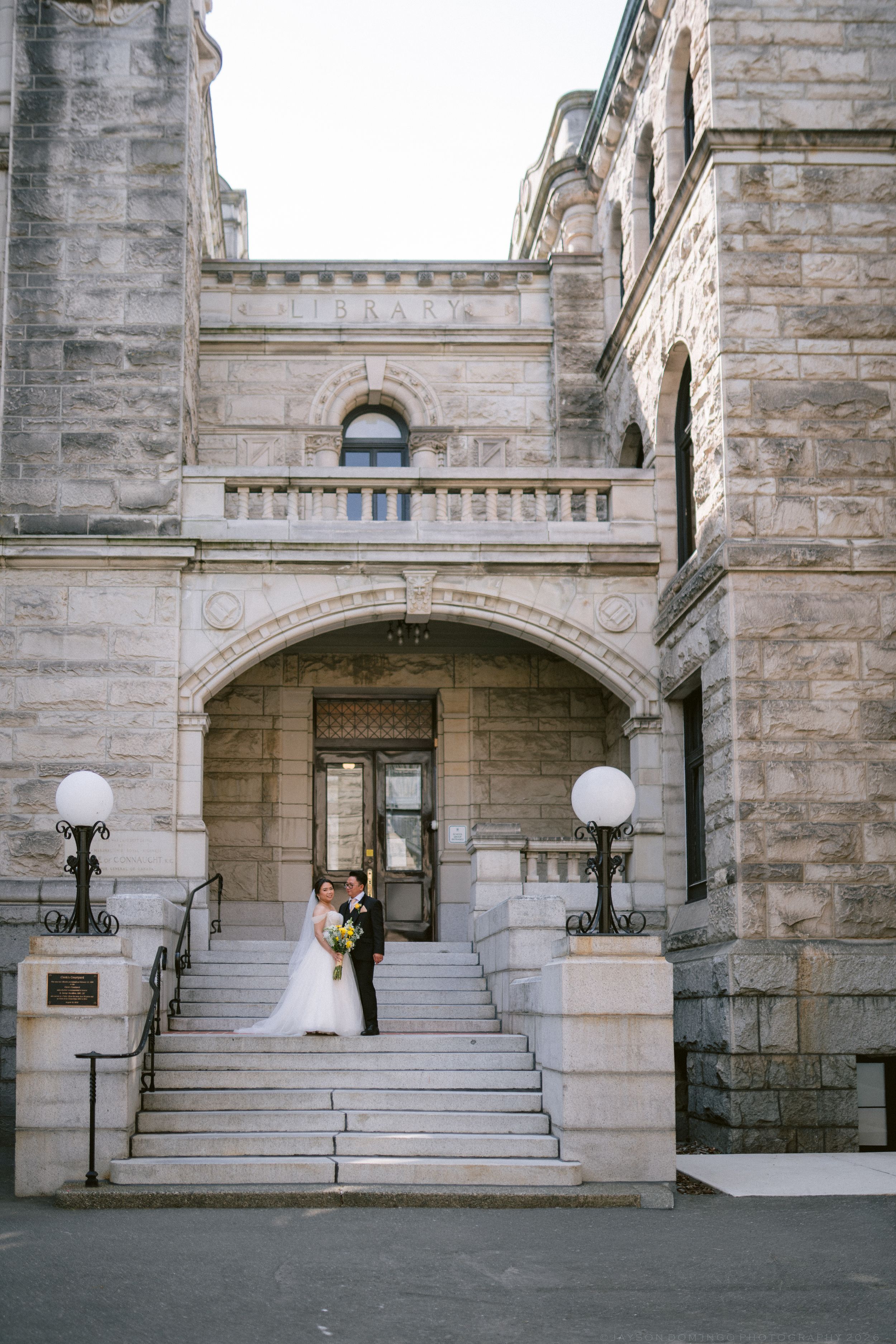 Bride and groom embracing on grand stone steps of historic library building with arched entrance and ornate architecture.