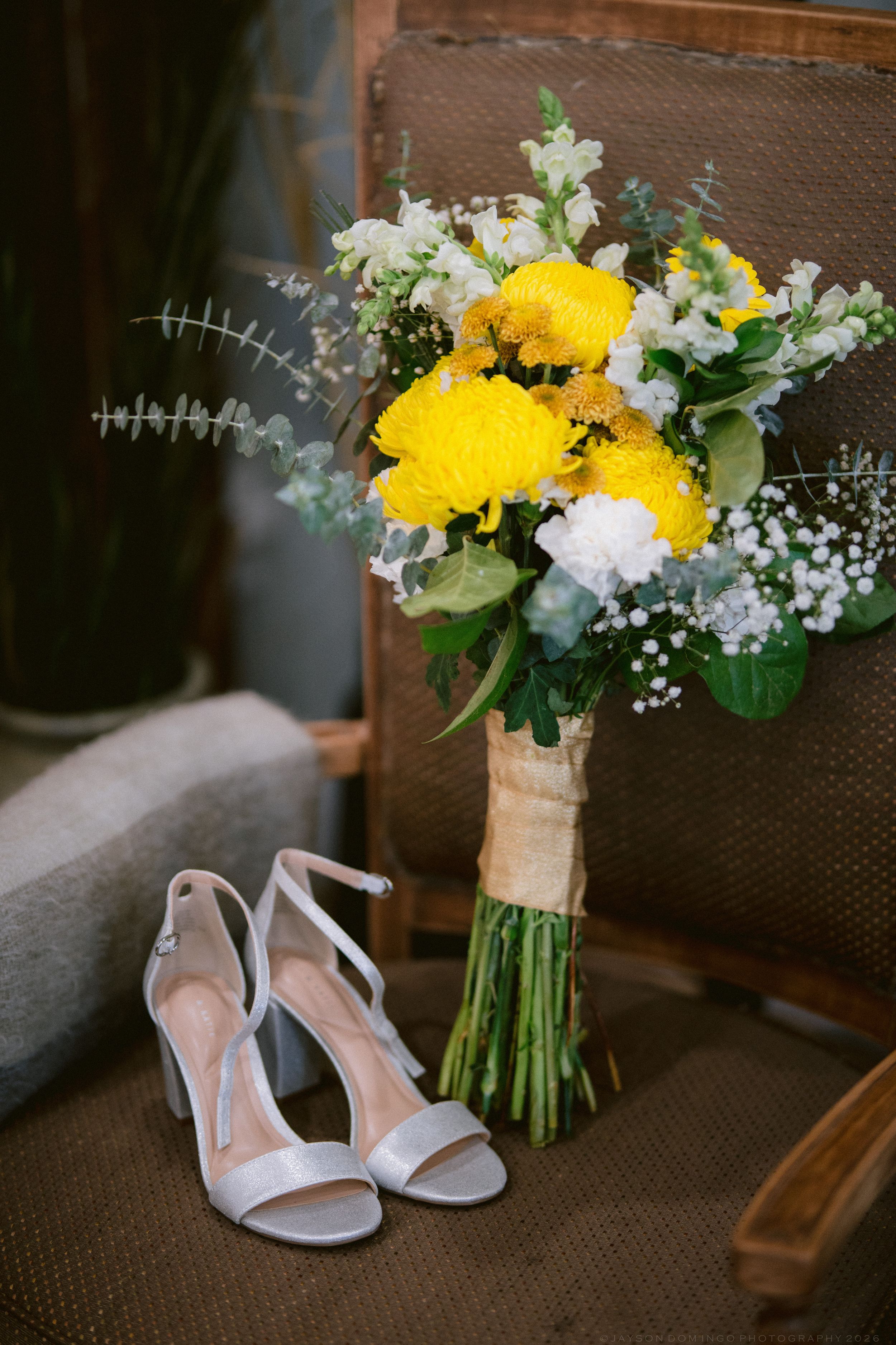 White bridal heels beside a yellow and white wedding bouquet with greenery on a wooden chair.