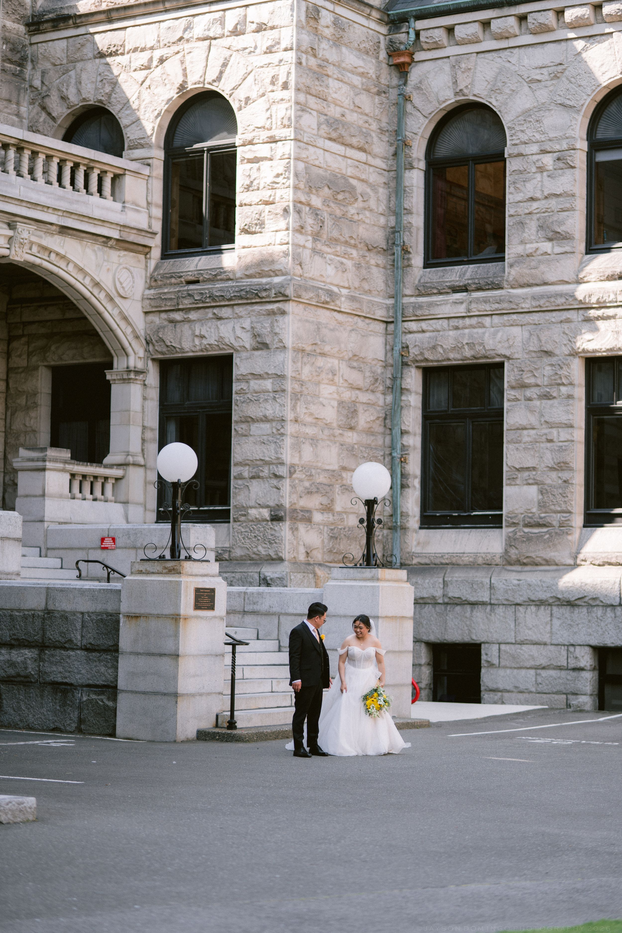 Bride and groom stand together outside grand stone wedding venue with arched windows and ornate architecture.