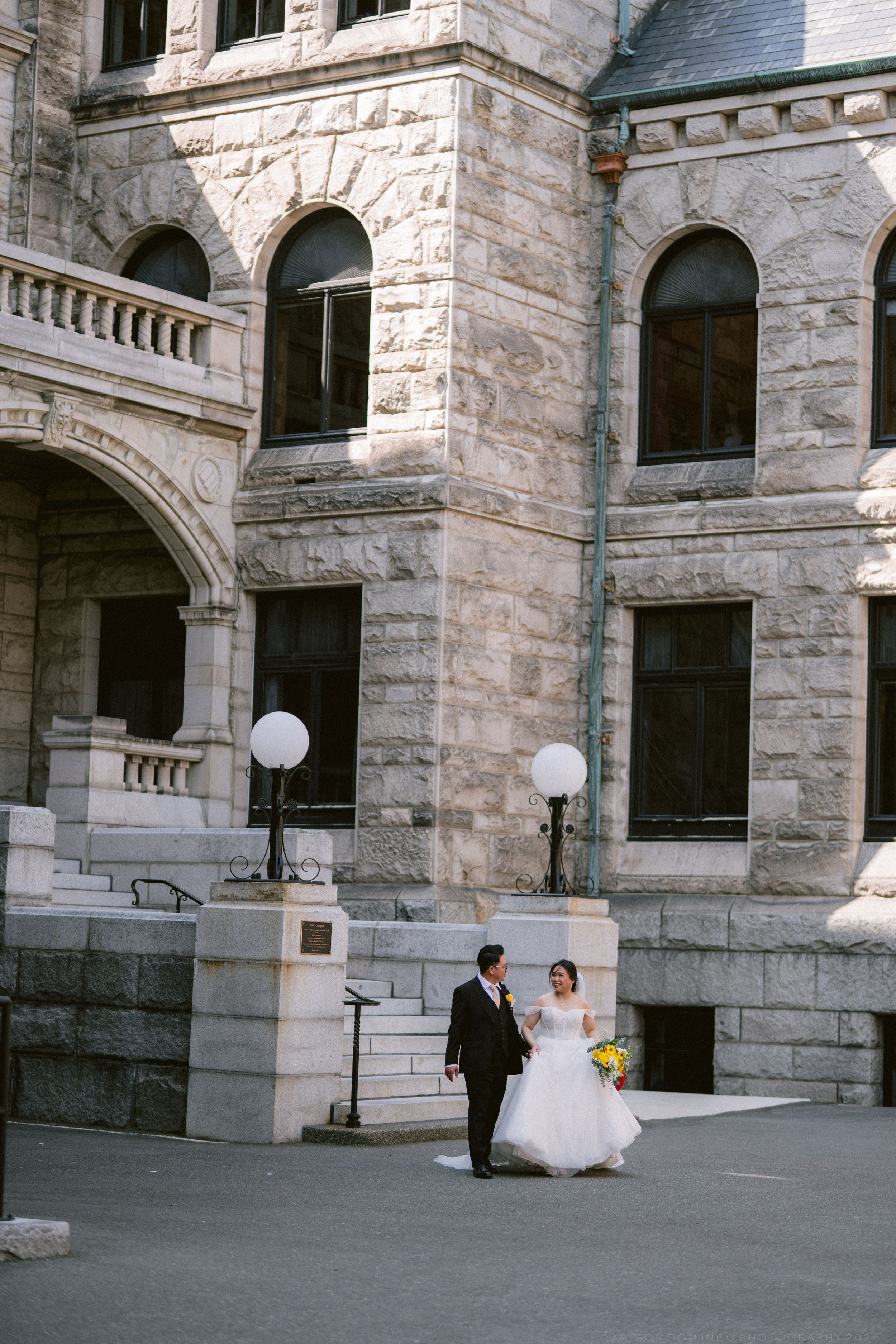 Bride and groom walk hand in hand outside a grand historic stone building on their wedding day.