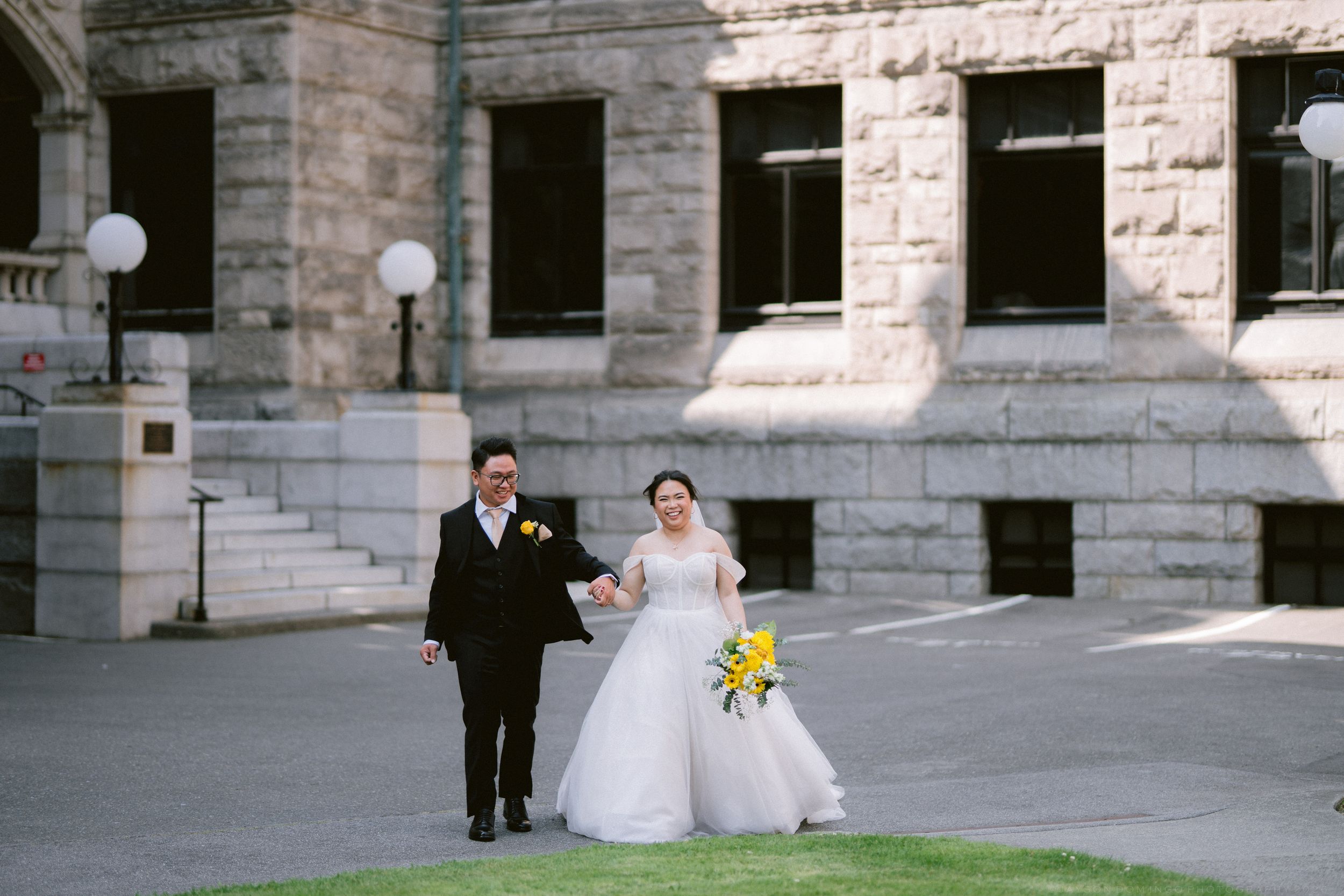 Bride and groom walk hand-in-hand across a sunny plaza in front of a historic stone building.
