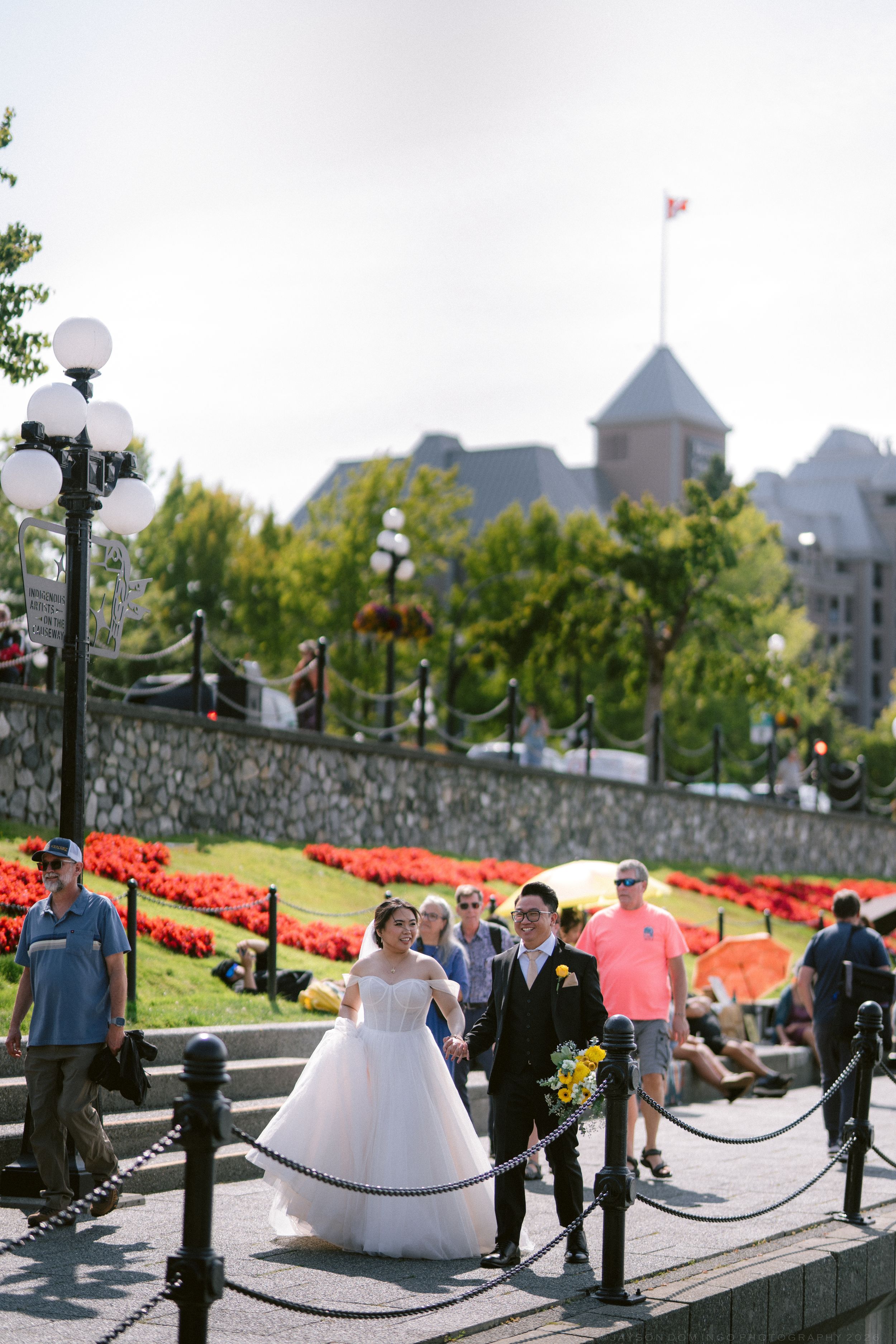 Bride and groom in formal attire walk hand in hand on outdoor steps with Canadian flag backdrop.