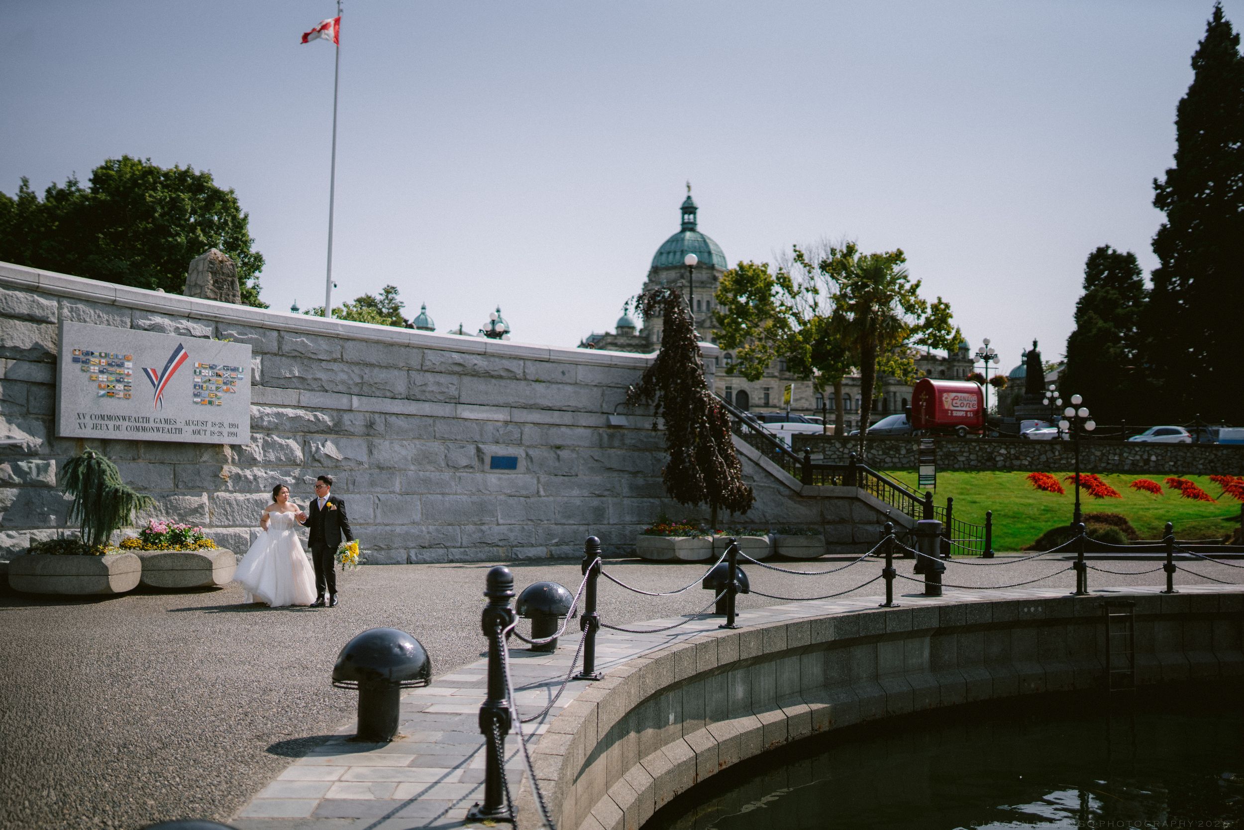 Bride and groom posing near waterfront plaza with Canadian flag and historic government building dome in background.