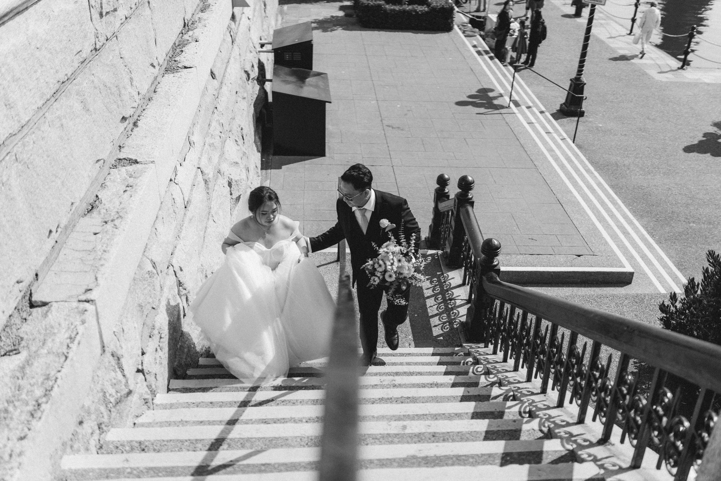 Black and white photo of a bride and groom walking up elegant outdoor stairs on their wedding day.