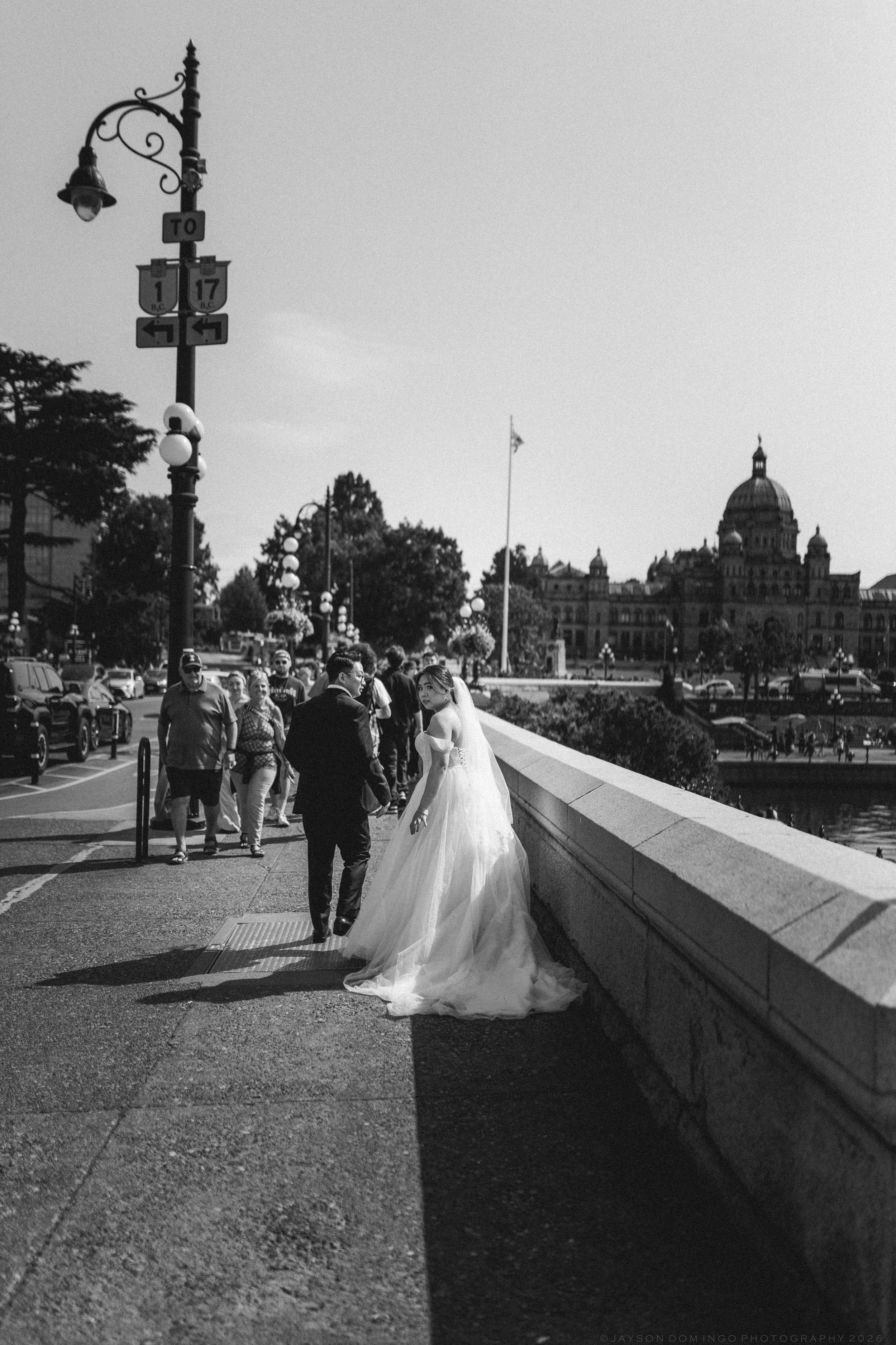Black and white photo of bride and groom walking along waterfront promenade near domed building.