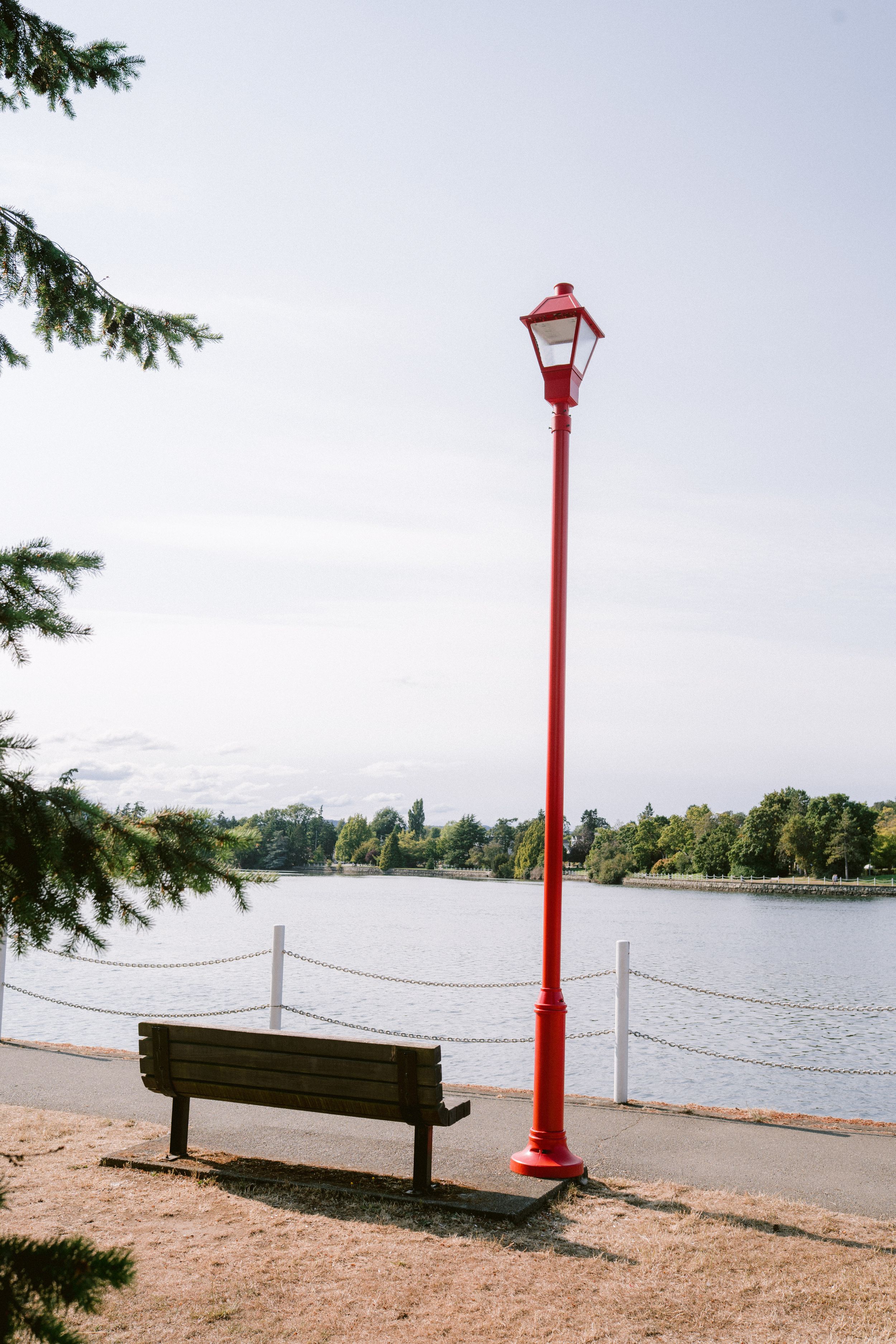 Red lamp post and wooden bench beside a calm lake in a scenic park setting.