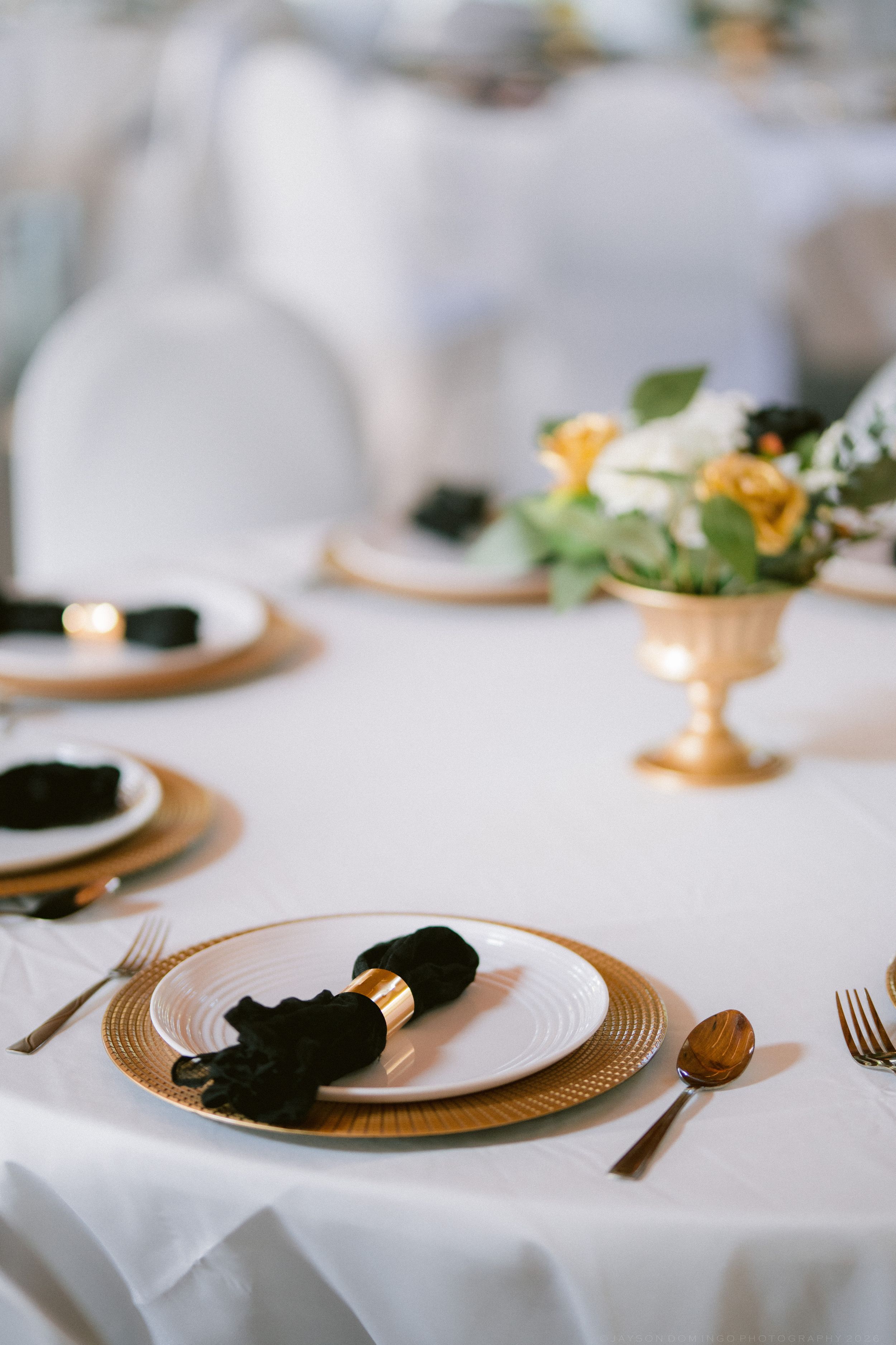 Elegant wedding reception table setting with gold chargers, black napkins, and yellow floral centerpiece.