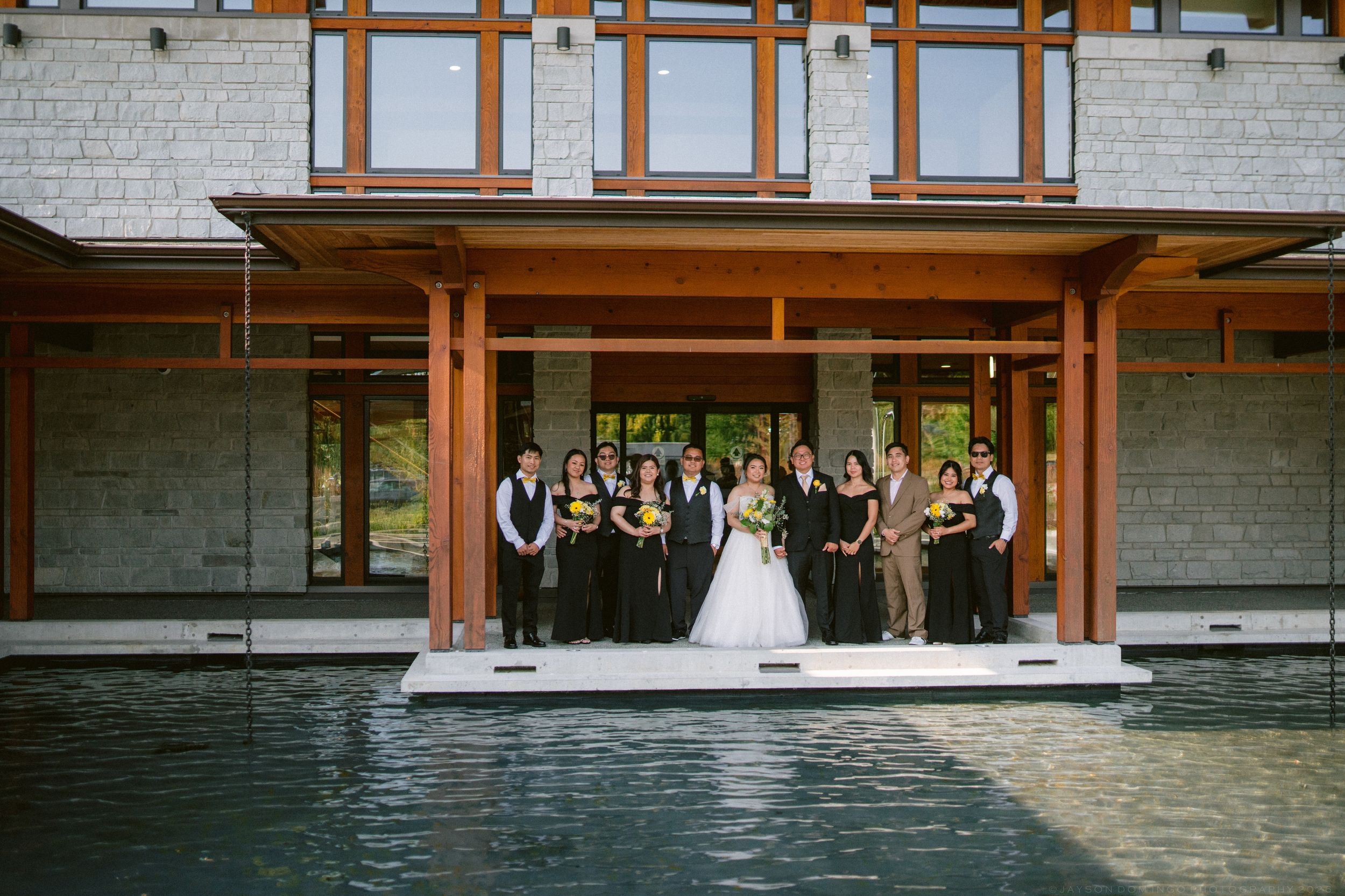Wedding party in black and tan attire poses together on a poolside deck of a modern wooden venue.