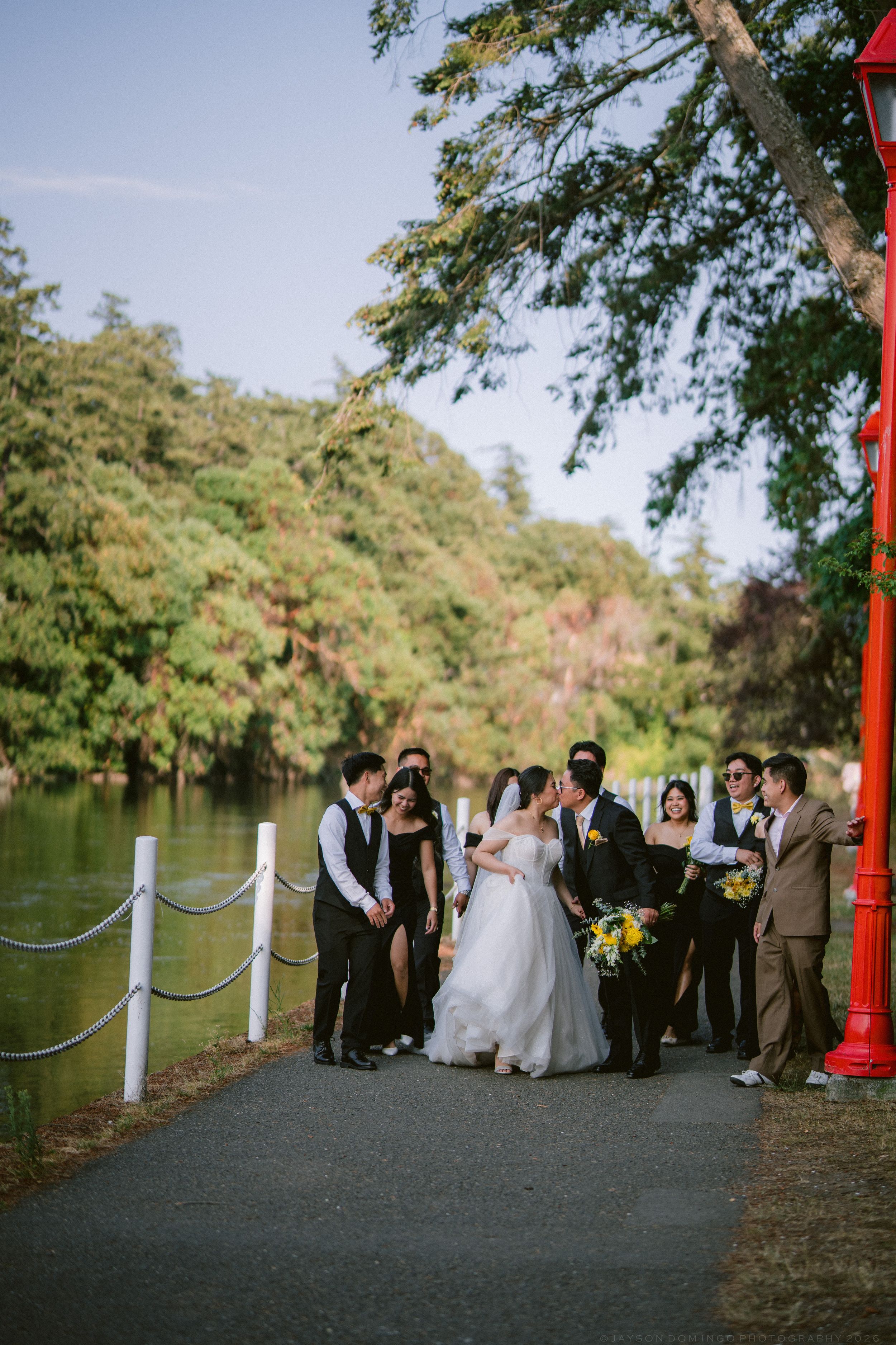 Wedding party gathered near lakeside red torii gate surrounded by lush green trees.