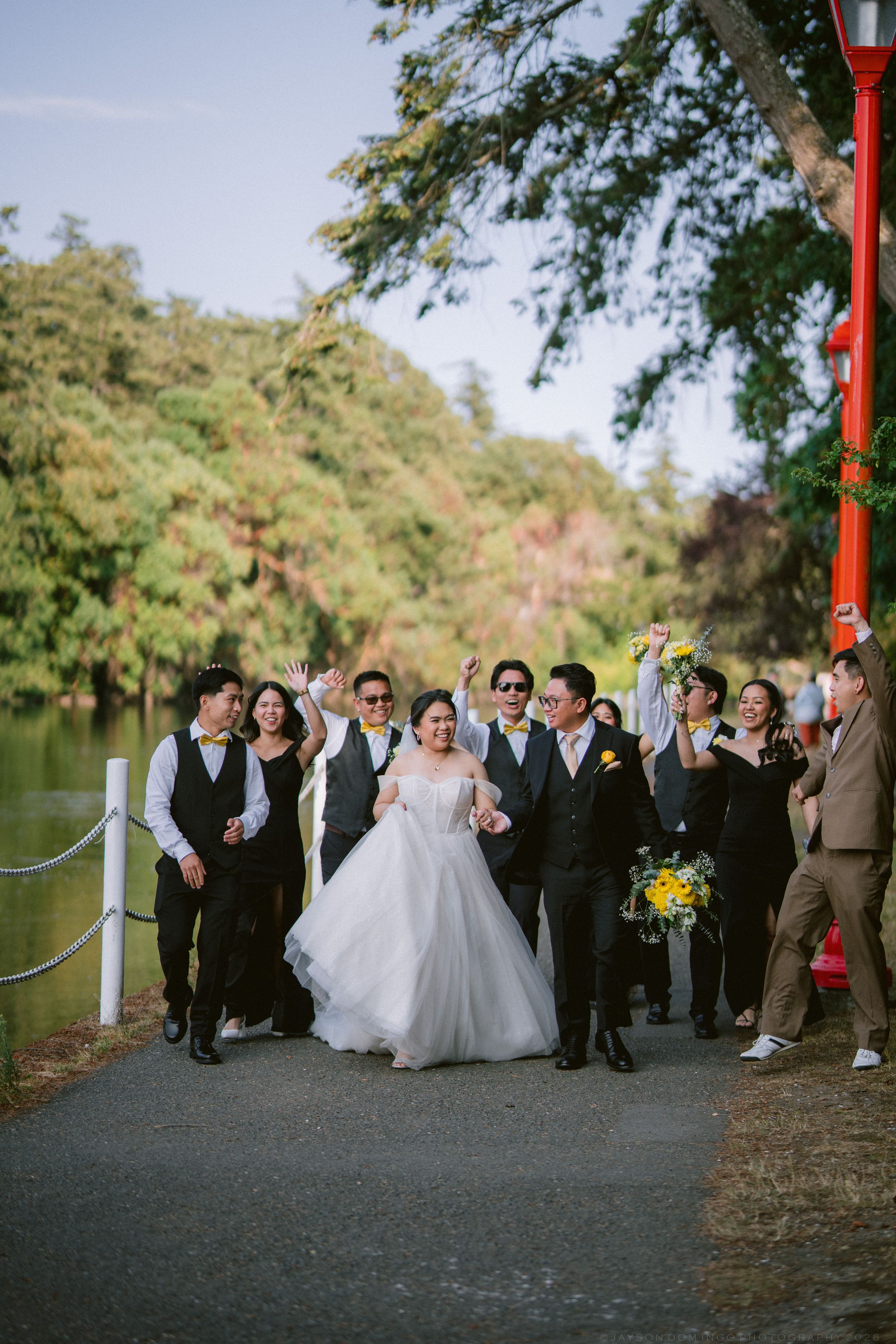 Joyful wedding party celebrating and walking along lakeside path near red torii gate.